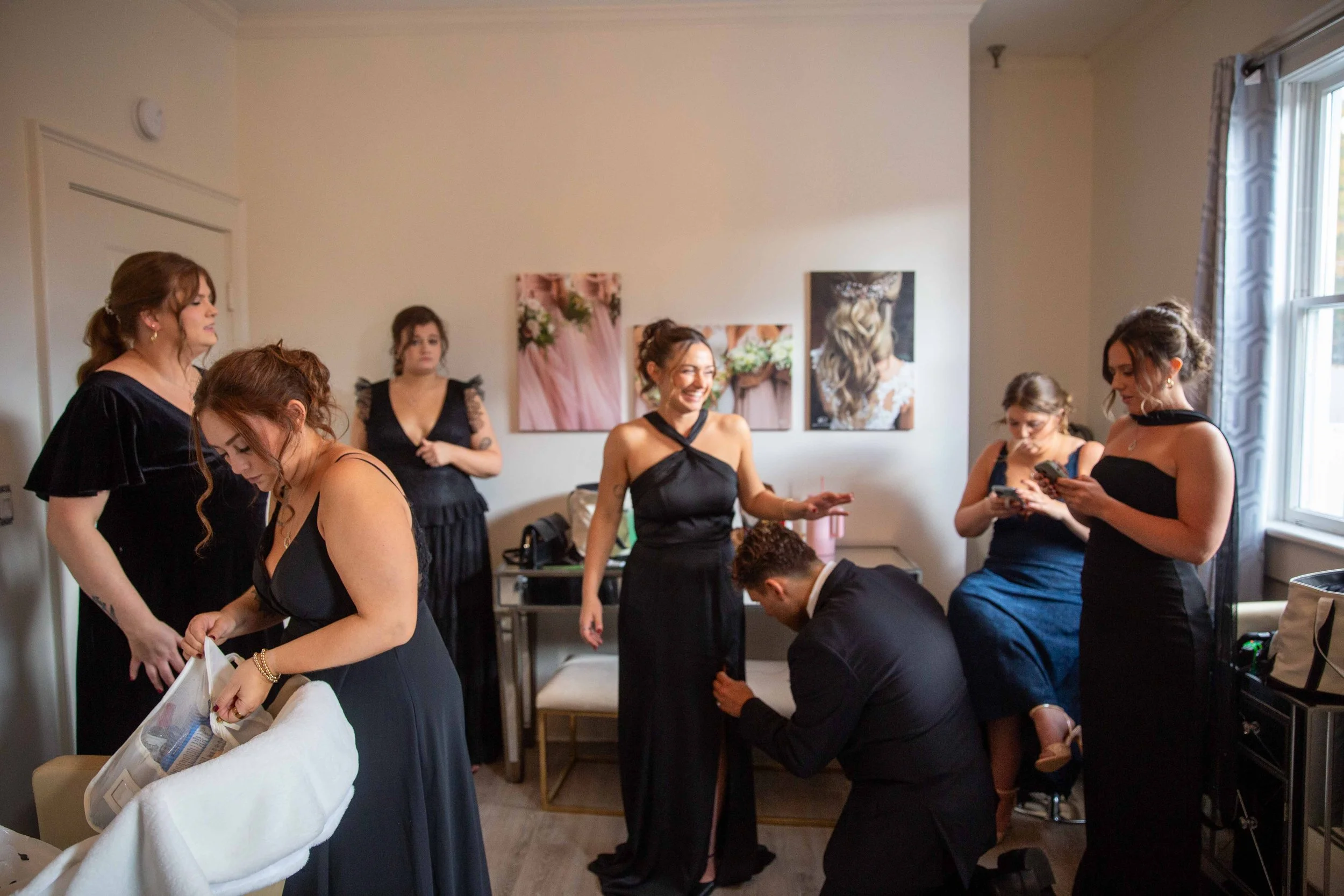 A group of women and a man preparing for a formal event in a well-lit room. Some women are on their phones, one woman is helping another with her dress, and a man is kneeling to adjust a woman's shoe or dress. The woman at the center is smiling. The 