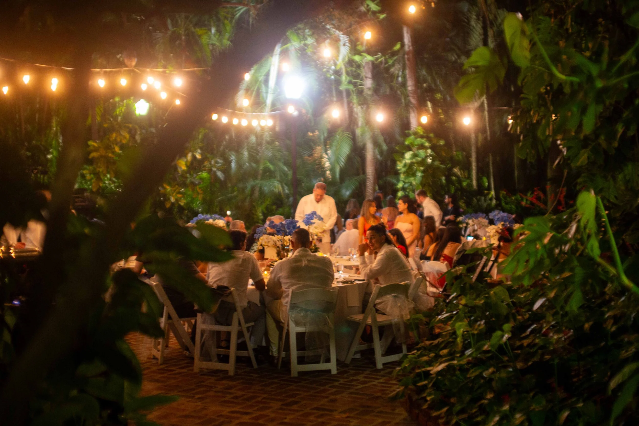 Guests dining at a long table decorated with floral centerpieces, outdoors at night with string lights overhead, surrounded by lush greenery.