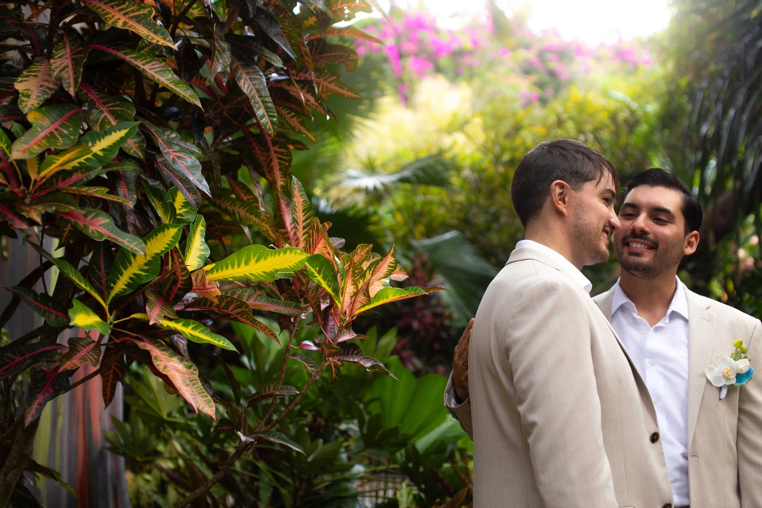 Two men dressed in light-colored suits, smiling happily, in a lush garden with colorful leaves and flowers in the background.