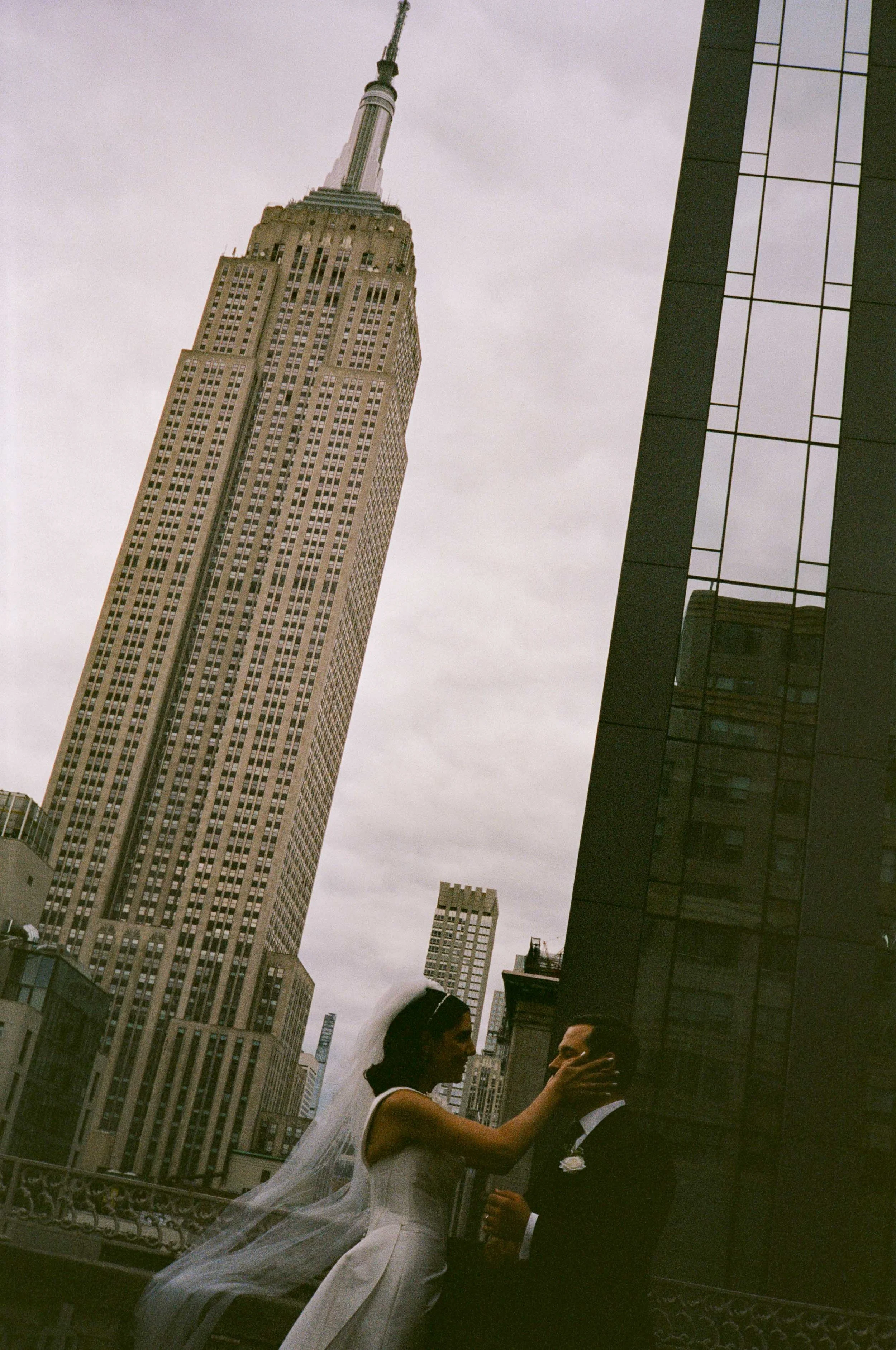 A bride and groom stand on a rooftop in New York City, with the Empire State Building in the background, during a cloudy day.