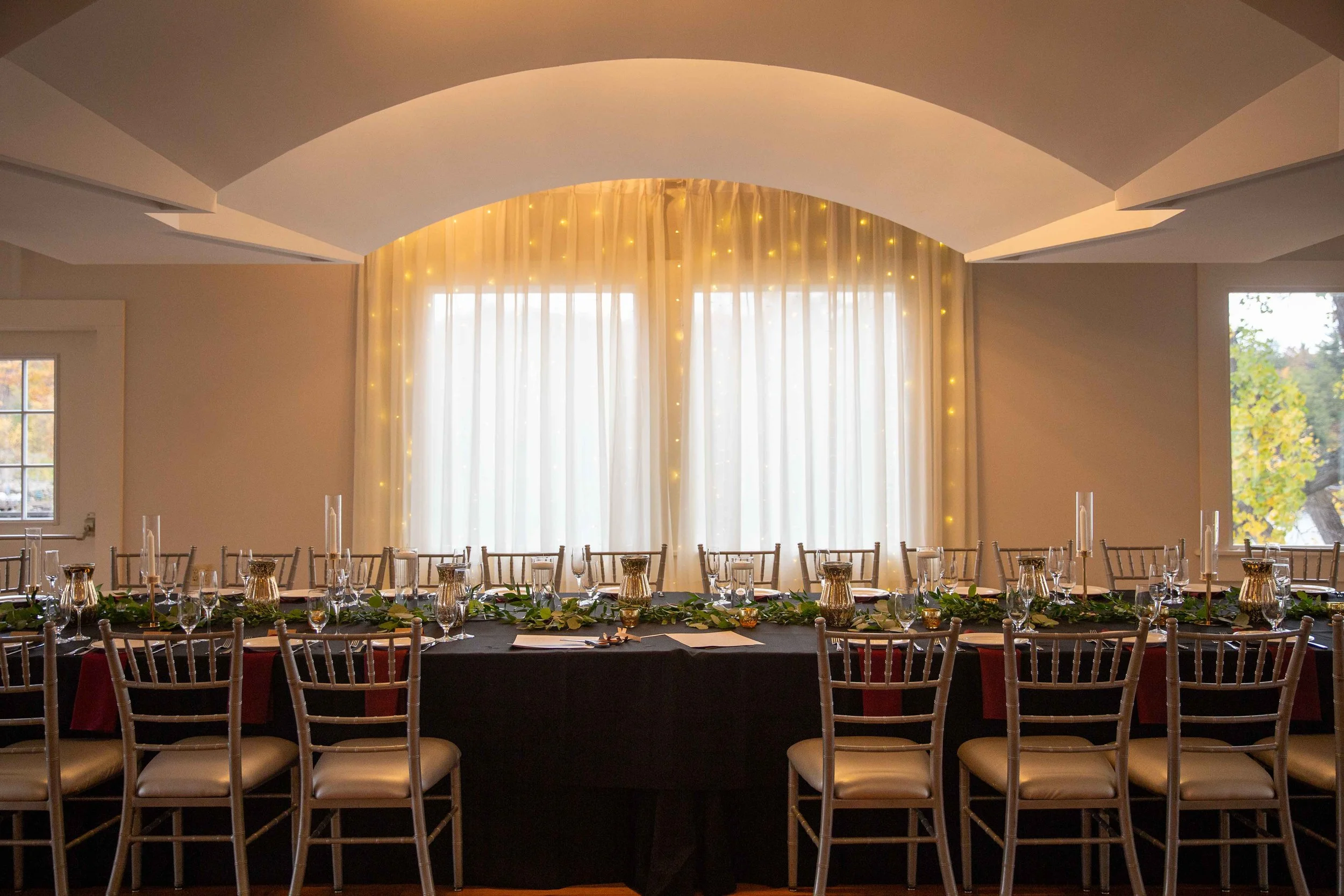 Long banquet table set for a formal event, decorated with candles, greenery, and gold accents, in a well-lit room with large windows and sheer white curtains.