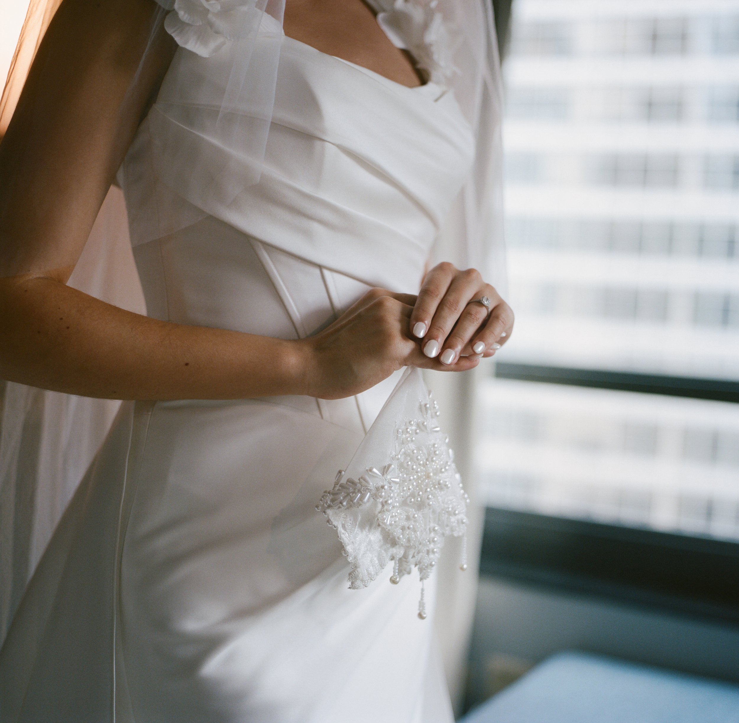 Close-up of a bride's hands holding a lace and beaded handkerchief, wearing a wedding dress with a fitted bodice and sheer sleeves, standing near a window with city buildings in the background.