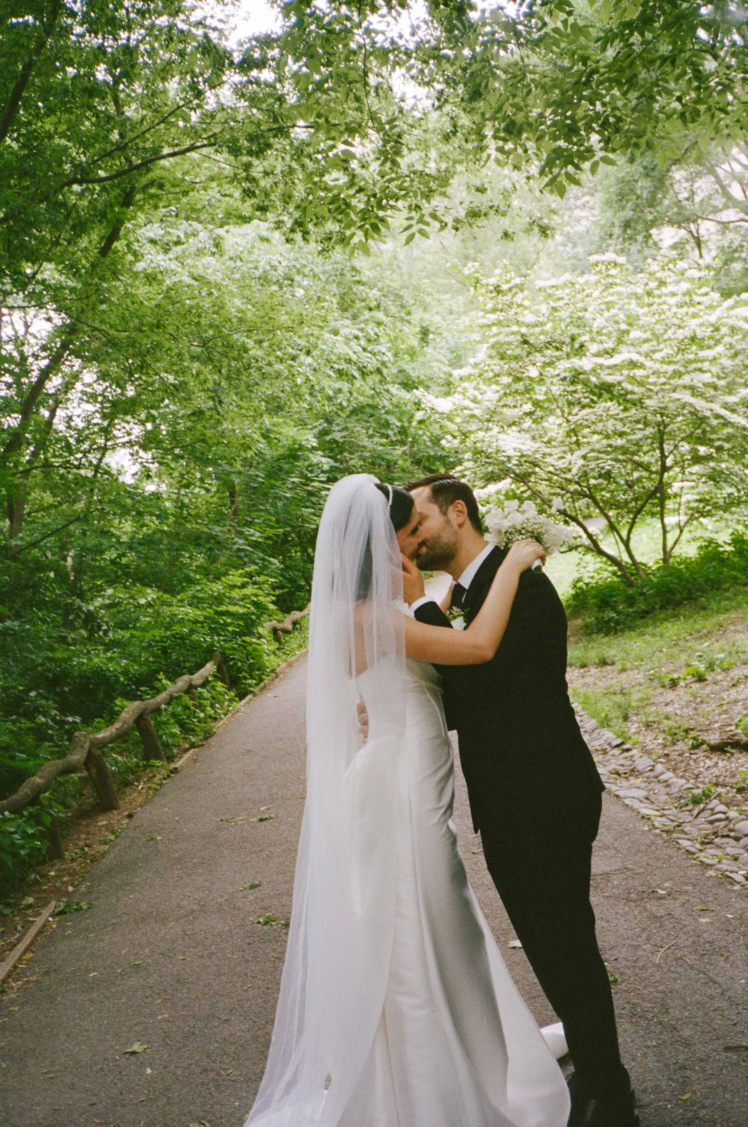 A bride and groom kissing on a forest path surrounded by lush green trees and foliage.