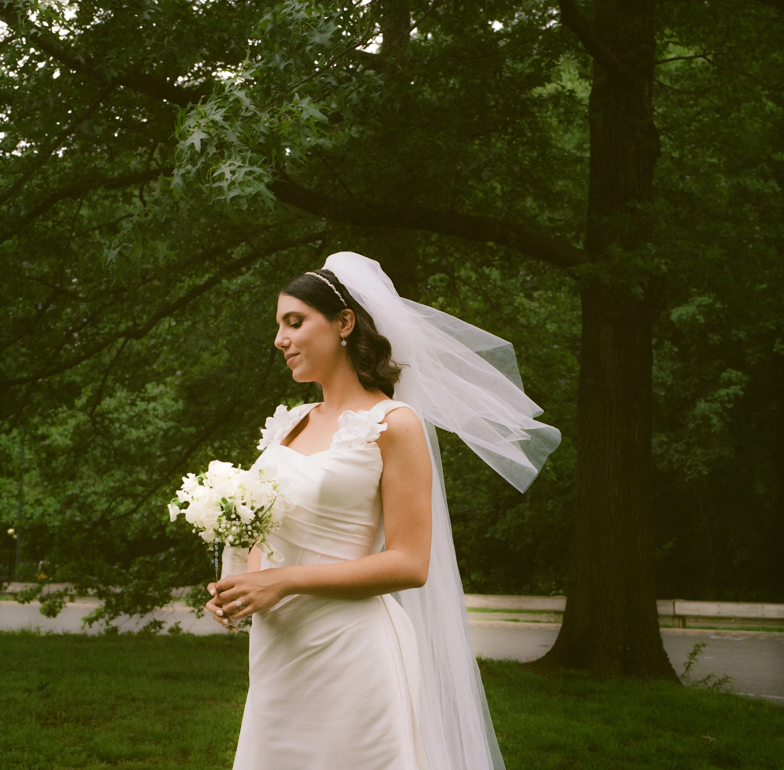 A bride in a white wedding dress holding a bouquet of white flowers stands outdoors in a park with green trees in the background.