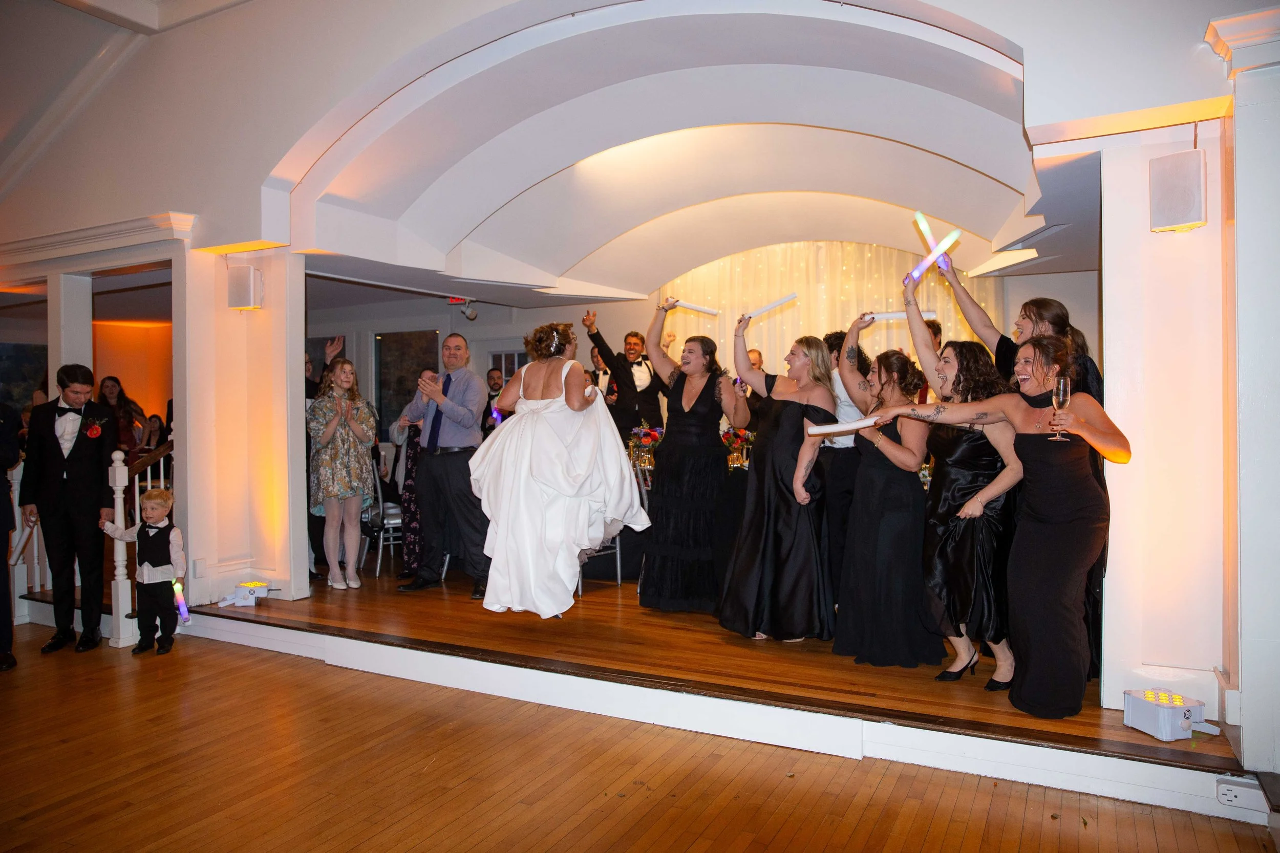 Wedding reception with bride in white dress and bridesmaids in black, celebrating with sparklers and glasses, guests clapping and watching.