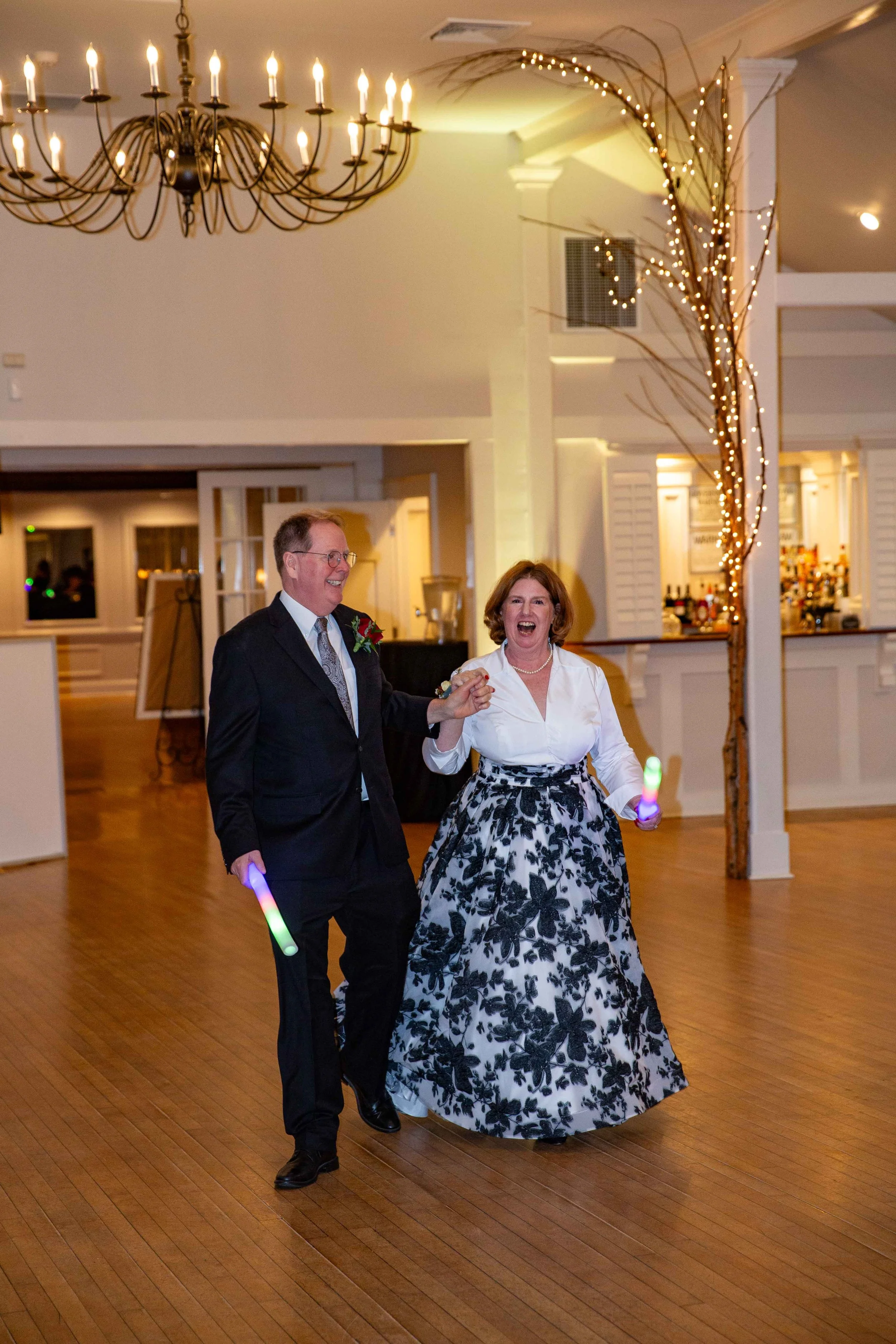 A man and woman dancing at a celebration, both holding glow sticks, in a decorated indoor venue with a chandelier and festive lighting.