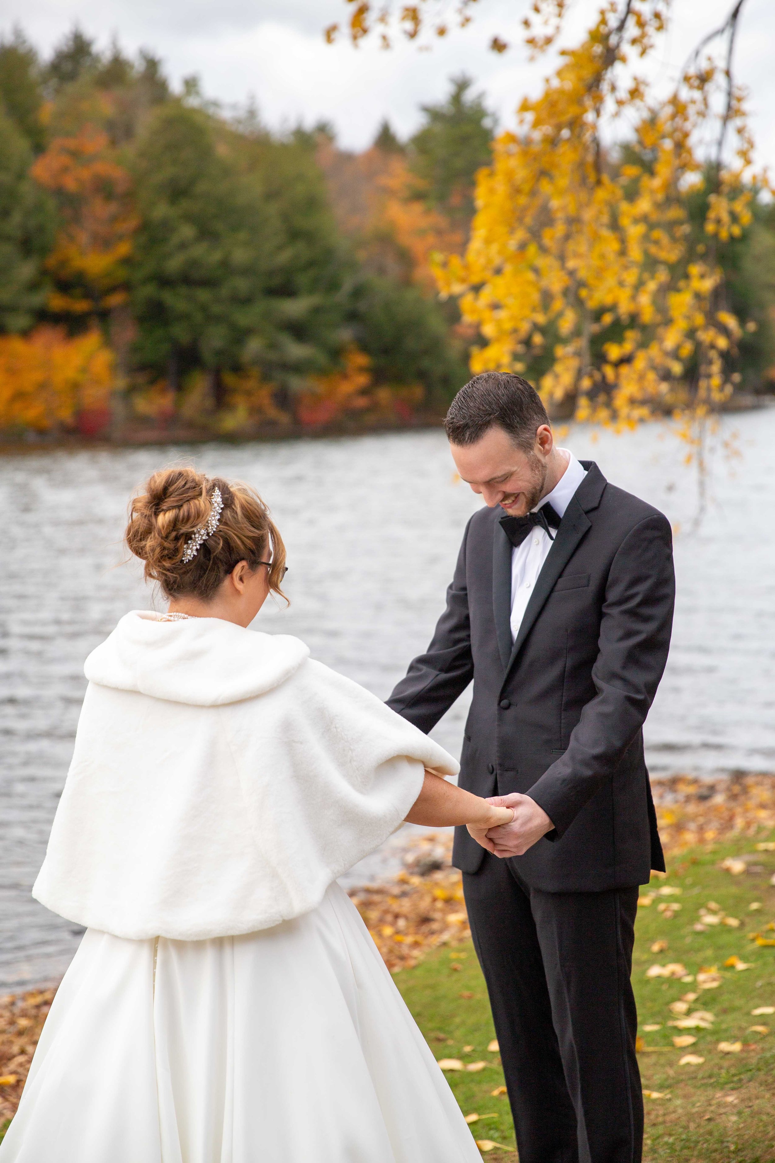 A bride and groom holding hands, standing by a lake surrounded by autumn trees, during their wedding.