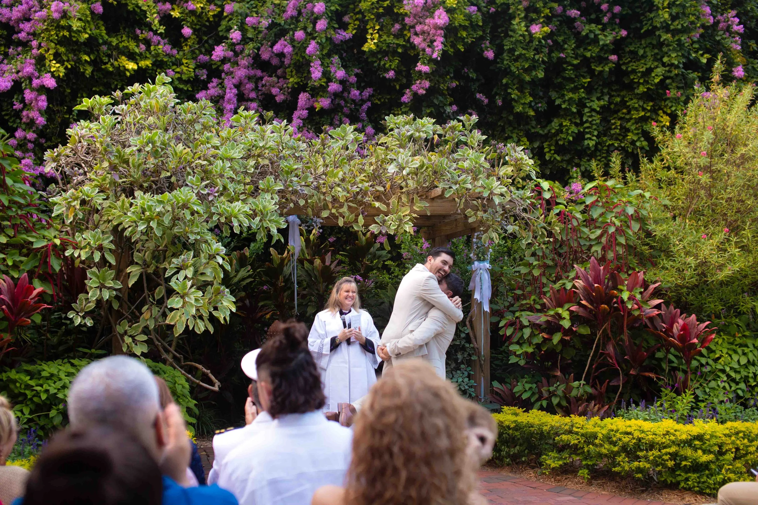 Just married couple embracing during wedding ceremony with officiant and guests in a lush garden setting with vibrant flowers and greenery.