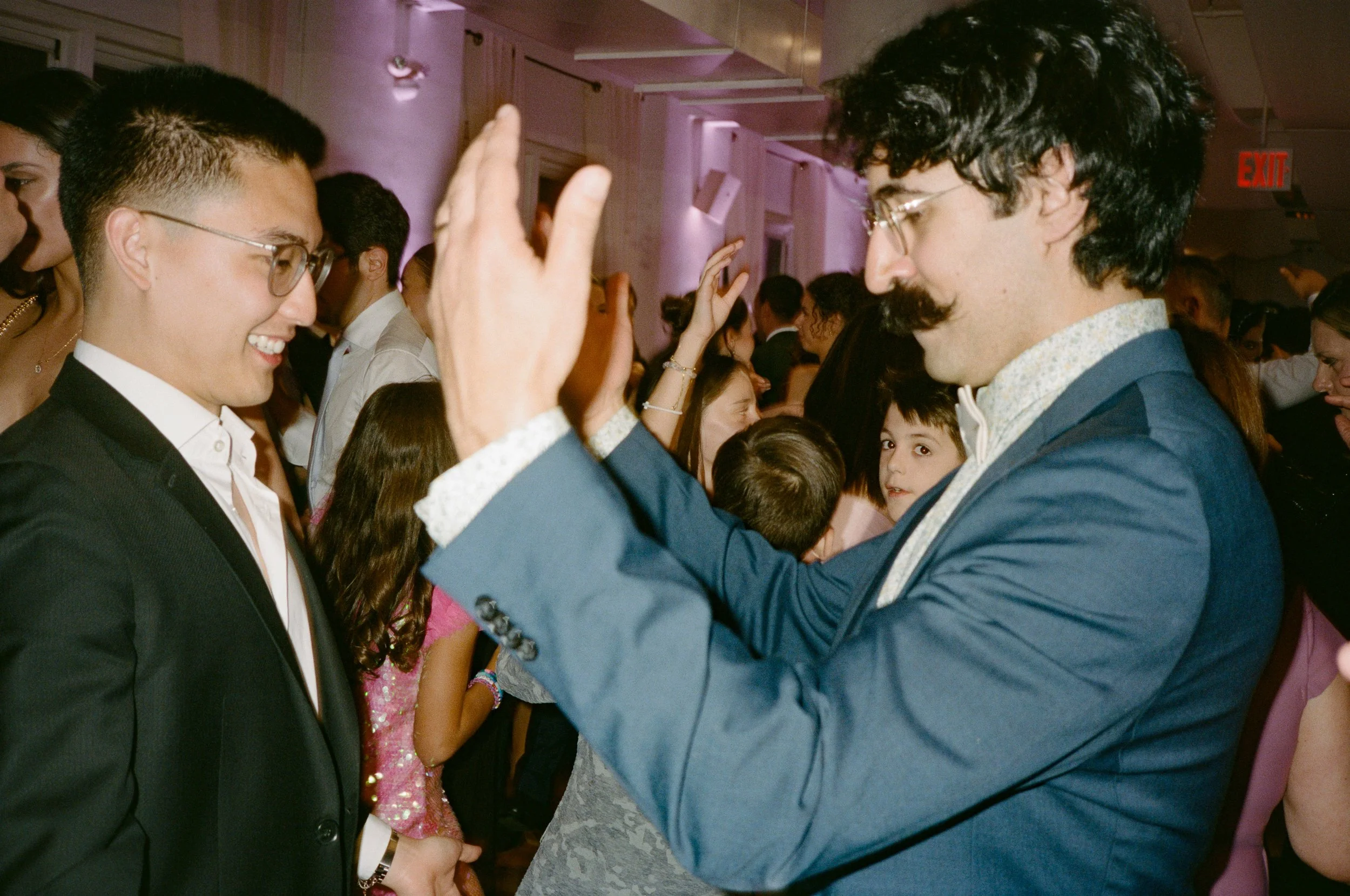 Two men in formal attire, one with glasses and a tuxedo, the other with a mustache and glasses, engaging in a friendly high-five at an indoor celebration or party with multiple people in the background.