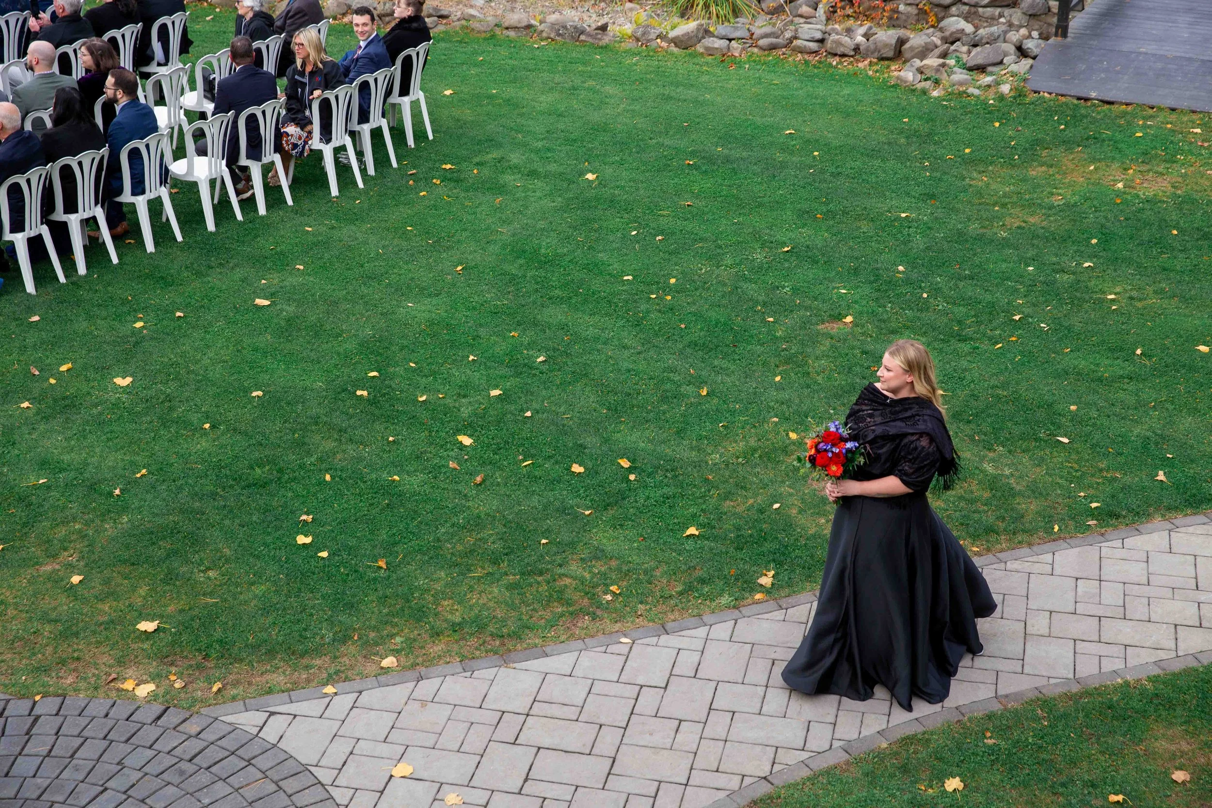 A woman dressed in a long black gown and shawl holding a bouquet of red, purple, and blue flowers walking on a paved path next to a green lawn with scattered yellow leaves, during an outdoor event with seated guests in the background.