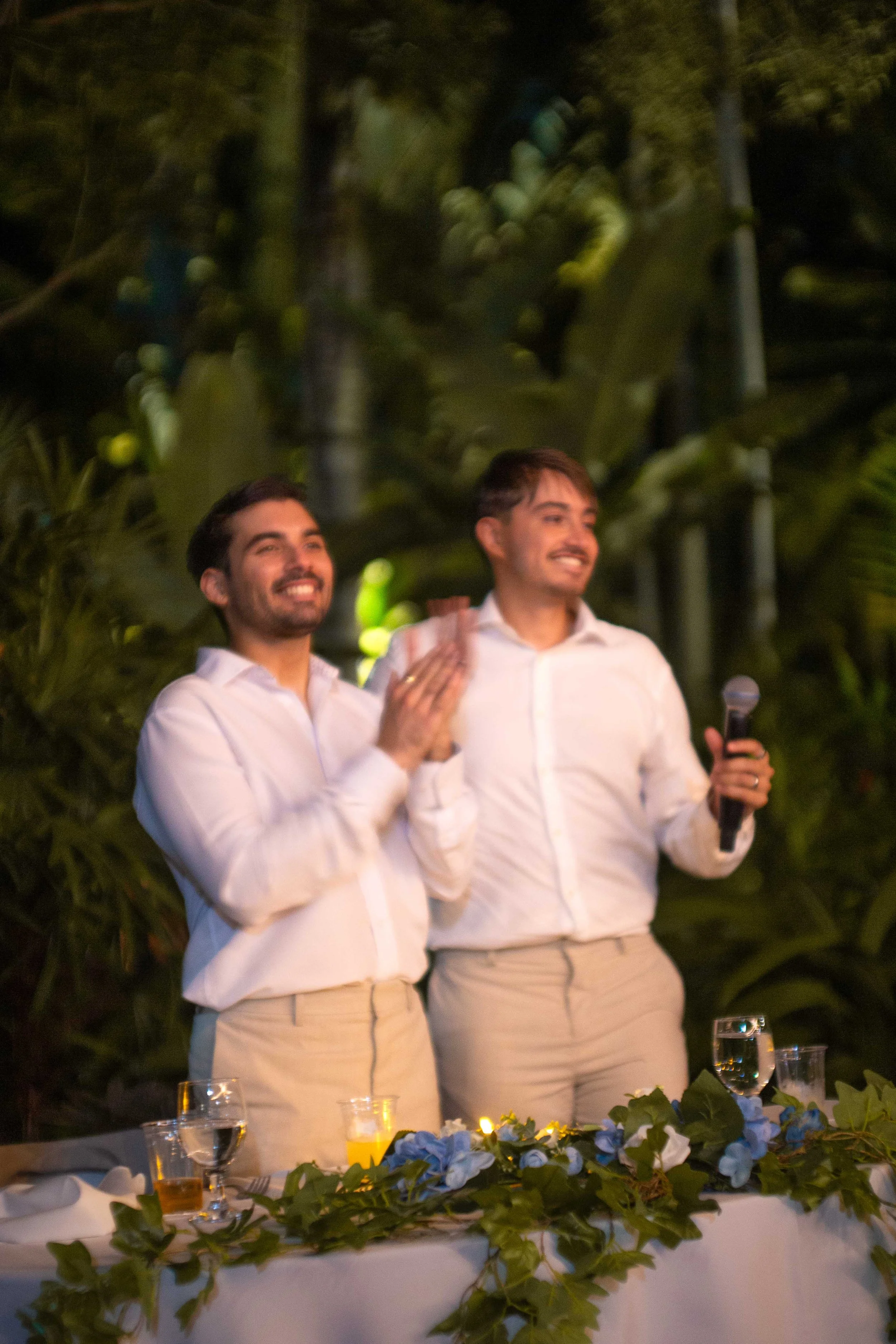 Two men in white shirts and beige pants standing behind a decorated table, smiling and celebrating at an outdoor event with greenery in the background. One man holds a microphone.