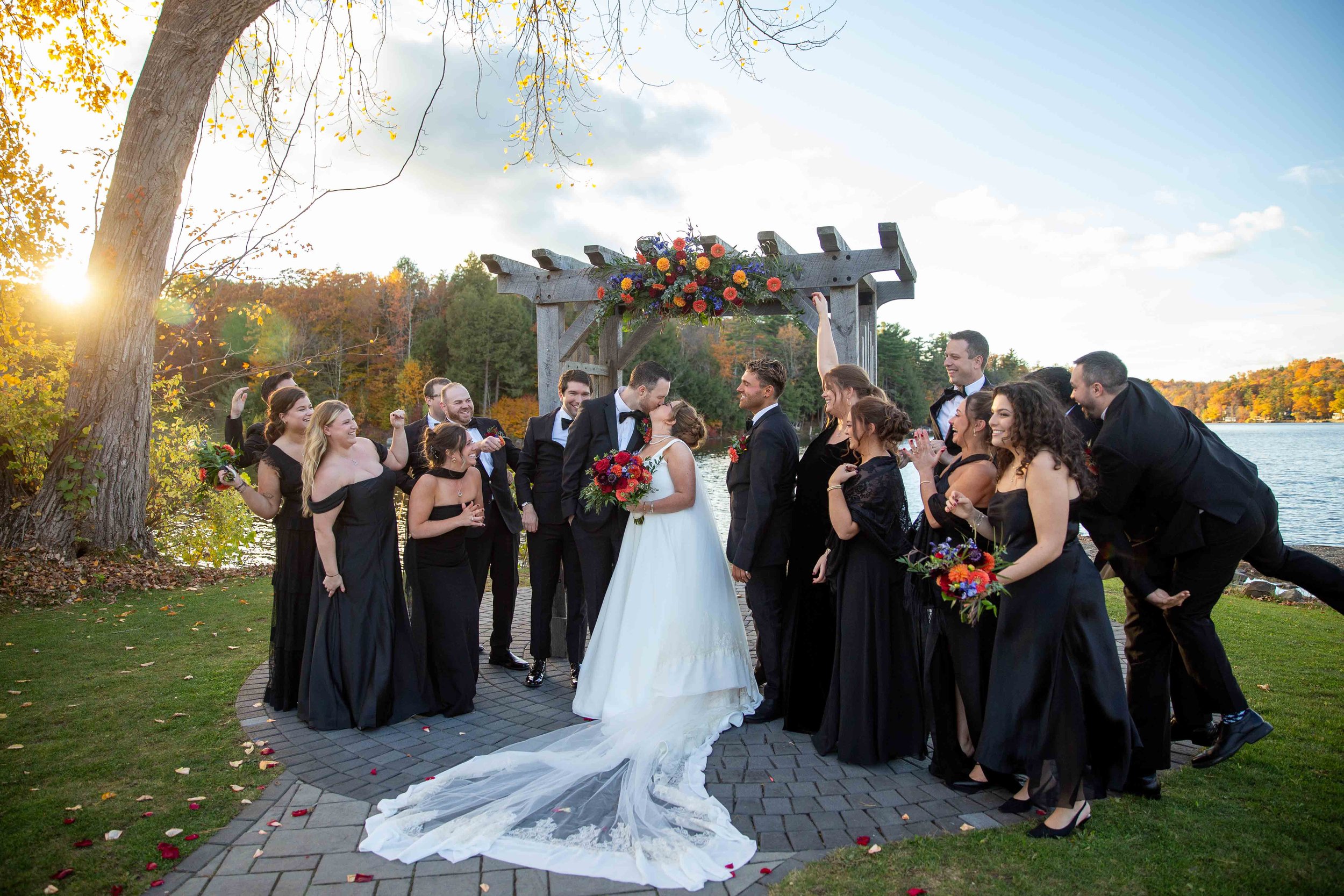 Wedding party including bride, groom, bridesmaids, and groomsmen celebrating outdoors near a lake at sunset, with fall foliage in the background.