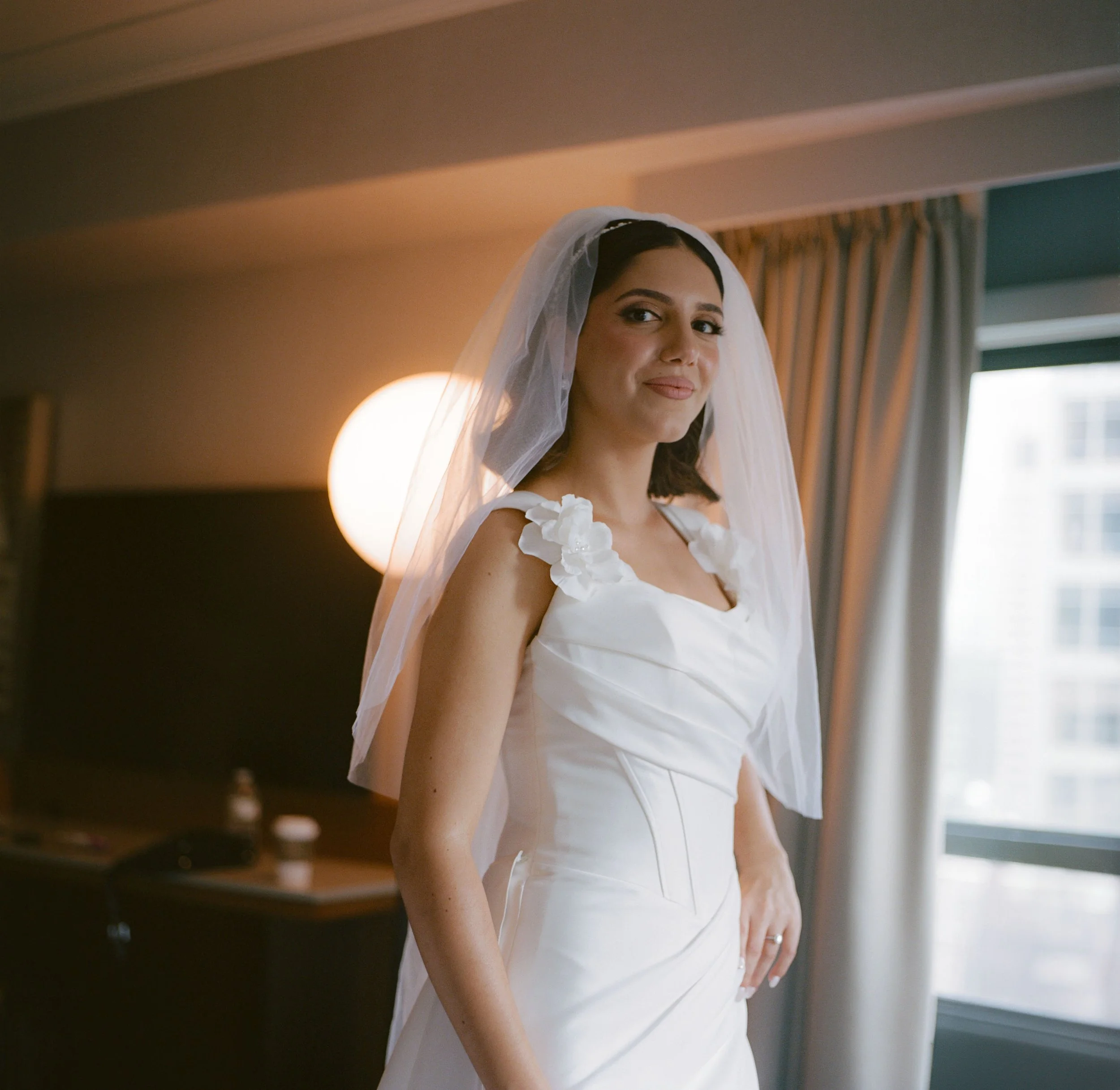 A bride in a white wedding dress with floral details on the straps, standing indoors near a window, with a veil on her head and a subtle smile.