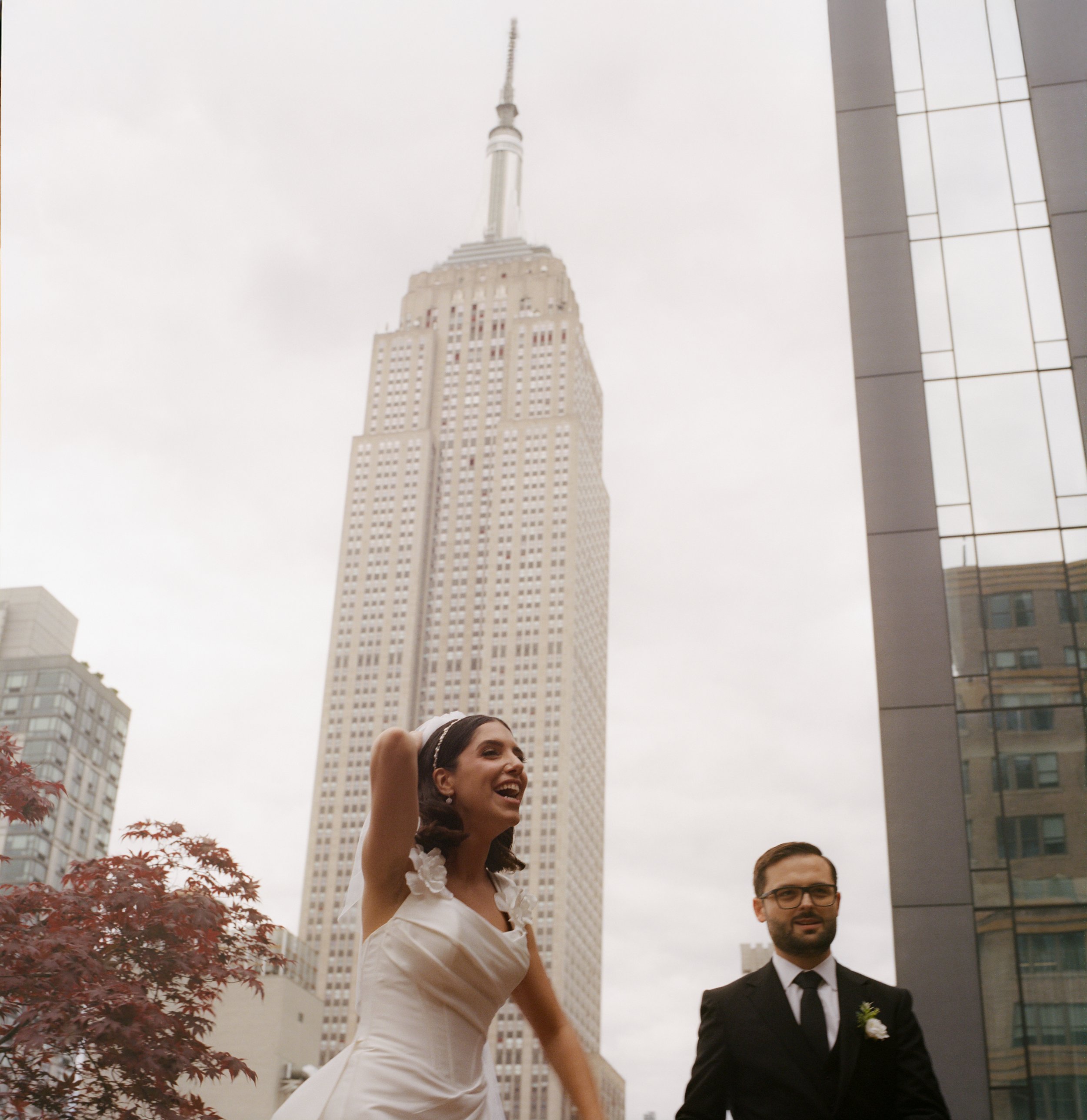 A woman in a white dress and a man in a black suit standing outdoors in front of the Empire State Building in New York City, with tall buildings and an overcast sky behind them.