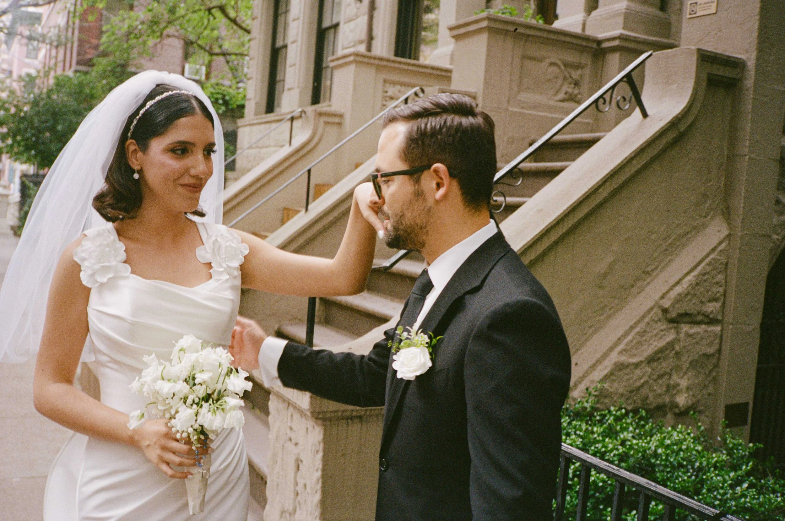 A bride in a white dress and veil holding a bouquet of white flowers gently touching the groom's forehead, who is wearing a black suit with a white shirt and glasses, standing outside on a city street with brownstone buildings in the background.