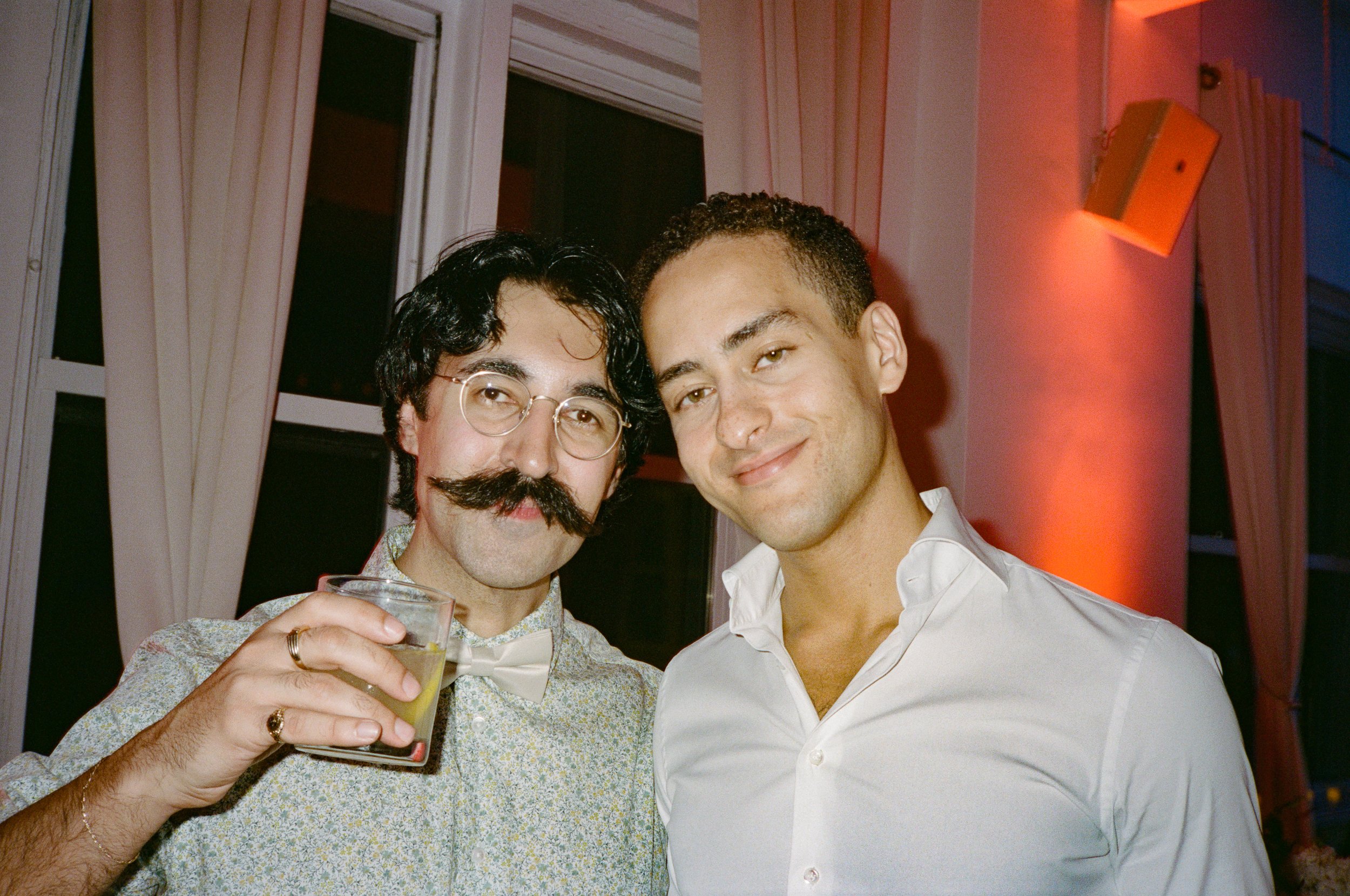 Two men standing close together, smiling at the camera in a warmly lit room with white and pink curtains and a window behind them. One man is holding a glass of a light-colored drink.