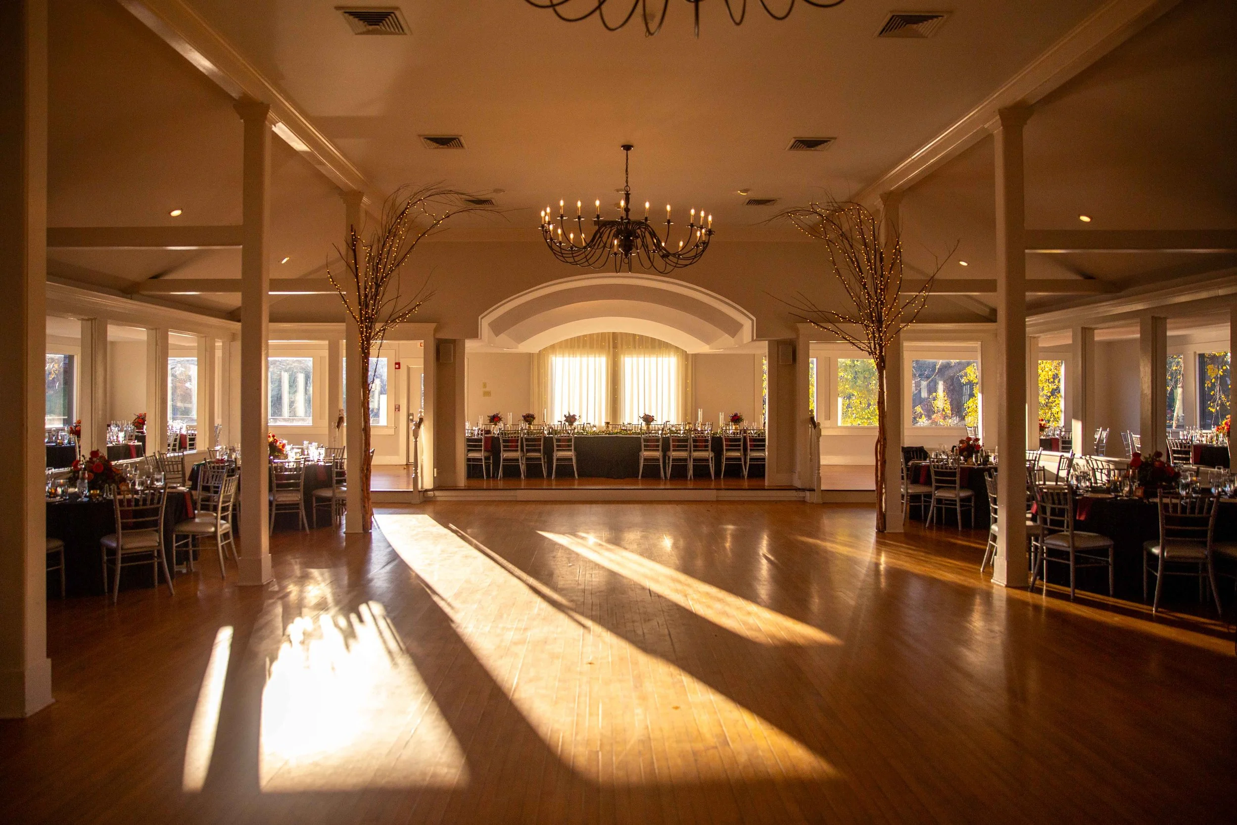 Empty banquet hall decorated for an event, with tables on the sides, a long head table at the far end, a chandelier hanging from the ceiling, and sunlight streaming through windows creating shadows on the wooden floor.