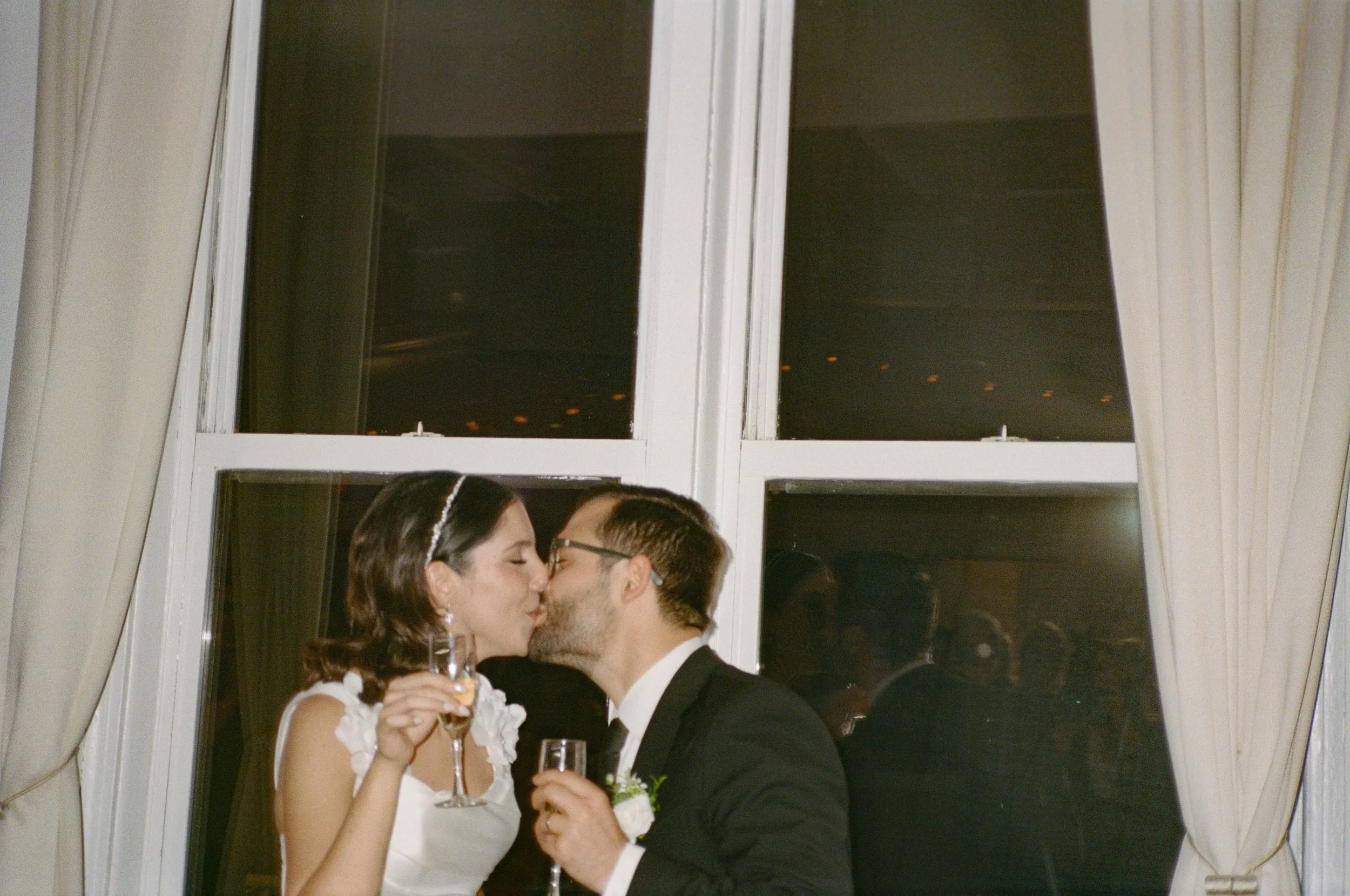 A bride and groom sharing a kiss at their wedding reception, holding champagne glasses, standing in front of a window with curtains.