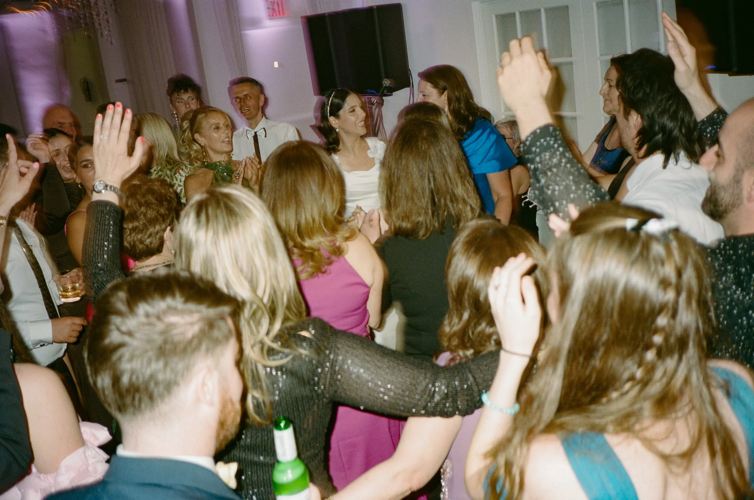 A crowded indoor party or celebration with people dancing, smiling, and socializing. Many have their hands raised, and some are holding drinks. The atmosphere appears lively and joyful.