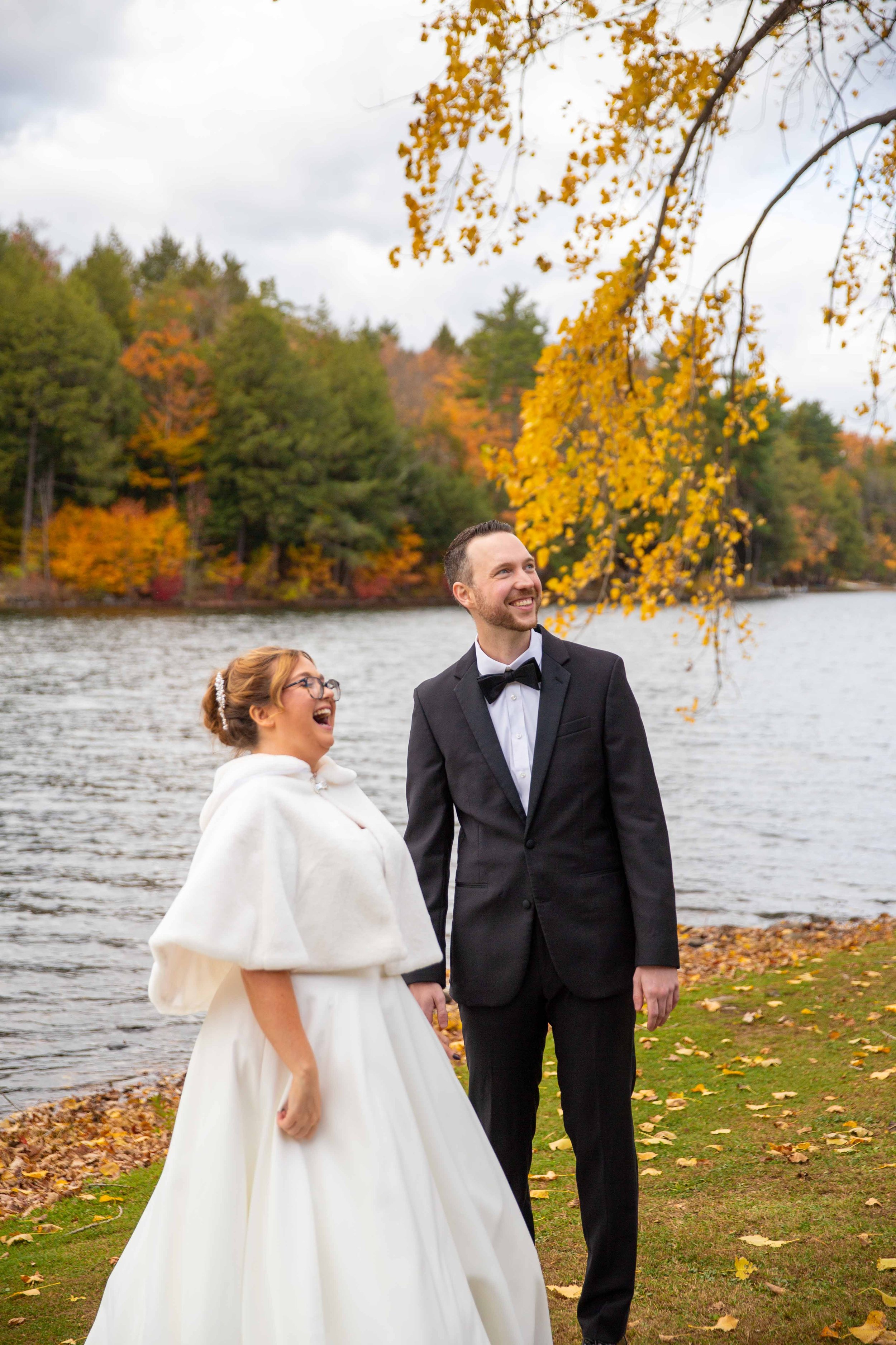 A newlywed couple stands outdoors near a lake, surrounded by colorful autumn trees, smiling and holding hands in wedding attire.