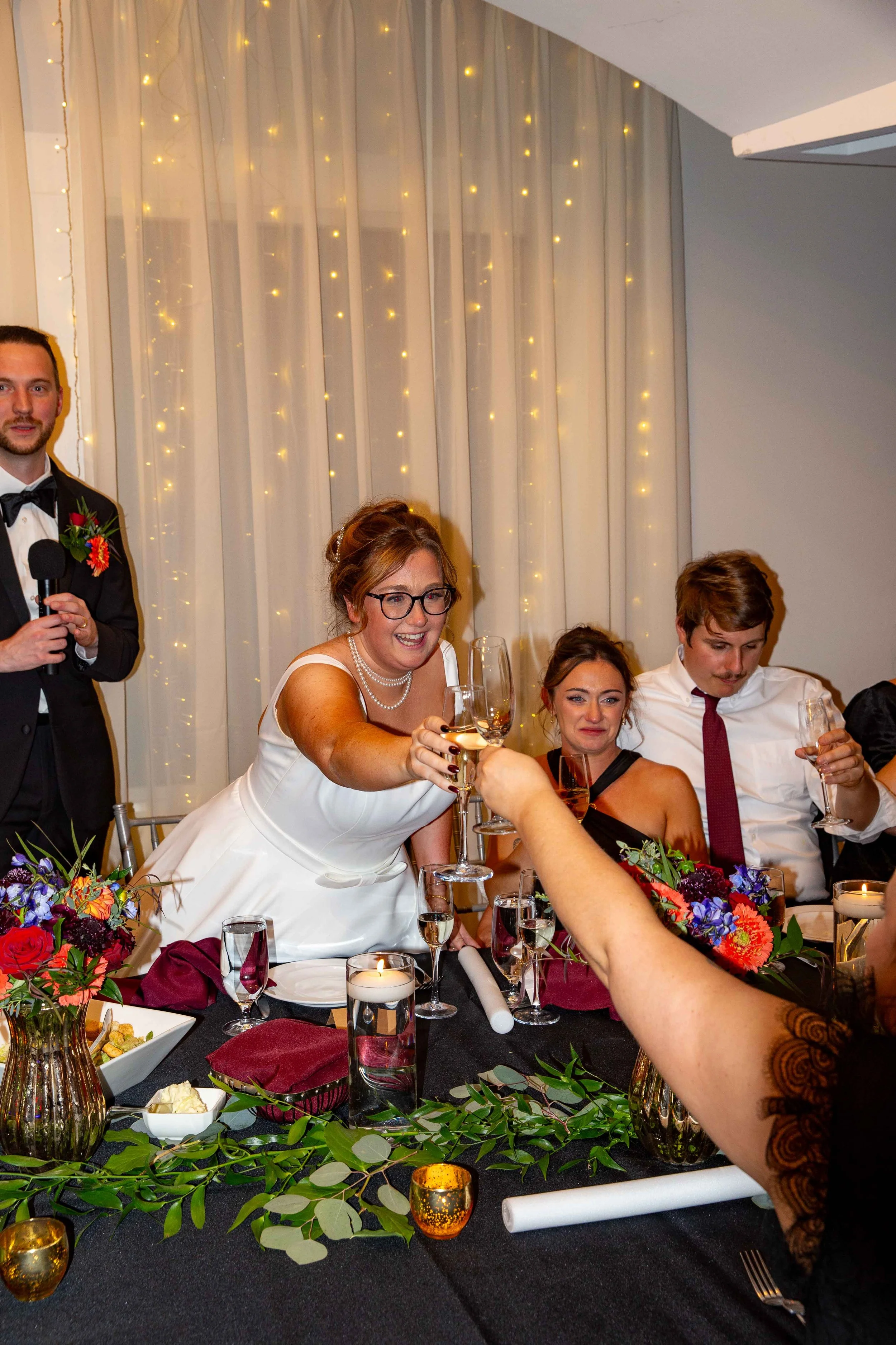Wedding reception with a bride in a white dress toasting with champagne along with seated guests, decorated with flowers, candles, and string lights.