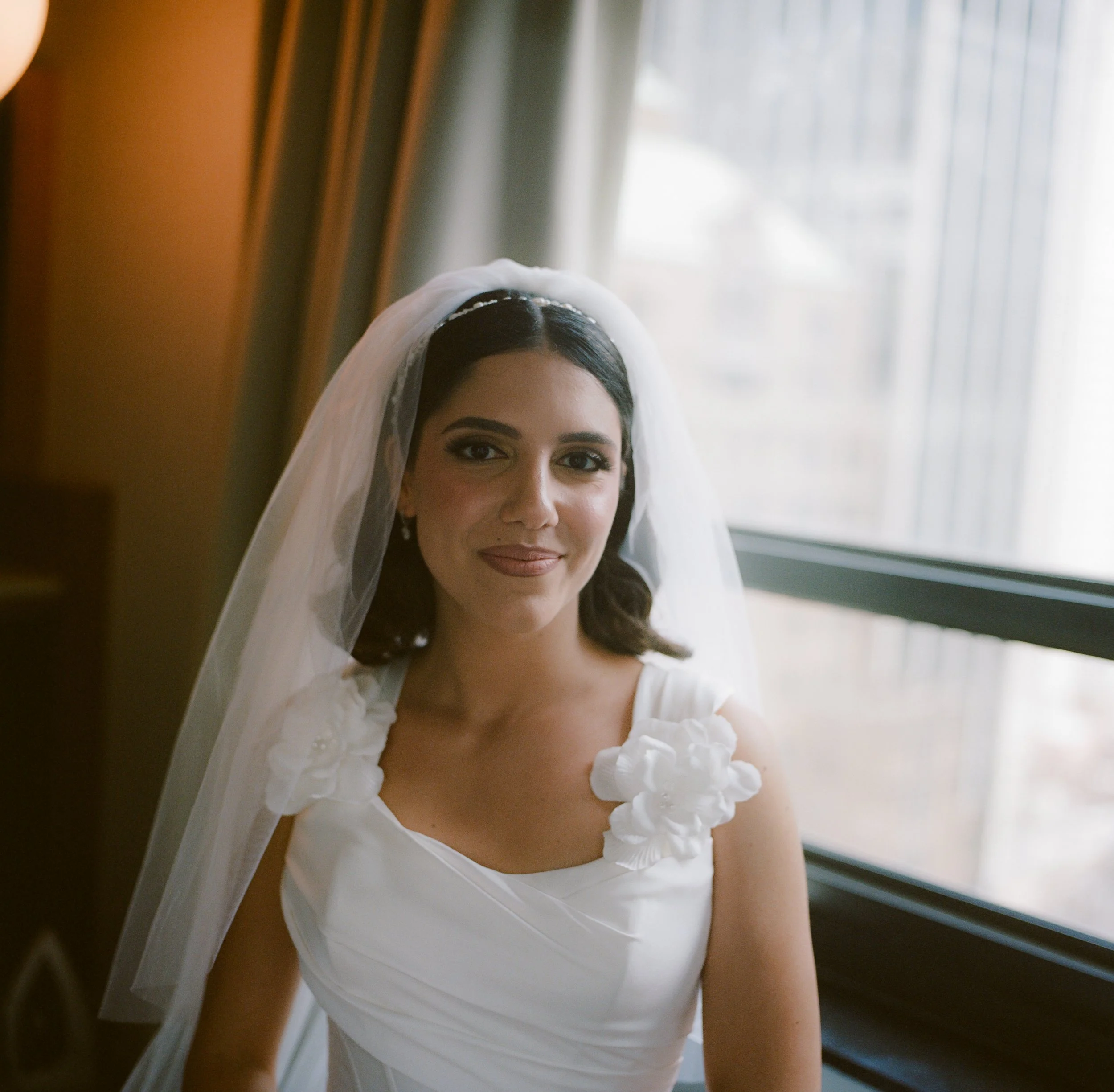 A bride with dark hair wearing a white wedding dress with floral accents on the shoulders, looking at the camera, standing next to a window with curtains.