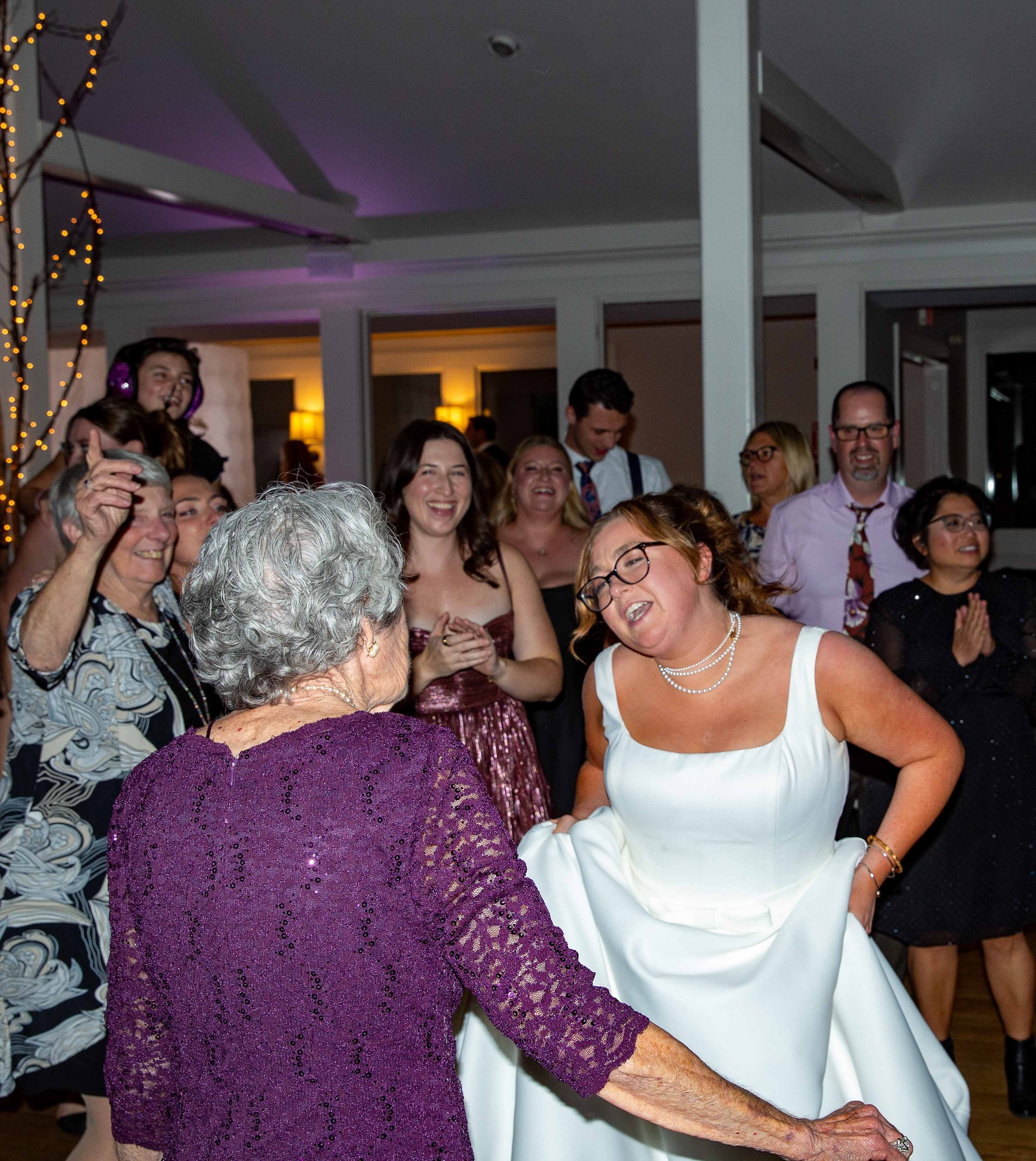 A woman in a white dress and glasses dancing with an elderly woman in a purple dress, surrounded by a group of people smiling and clapping at a celebration or party.
