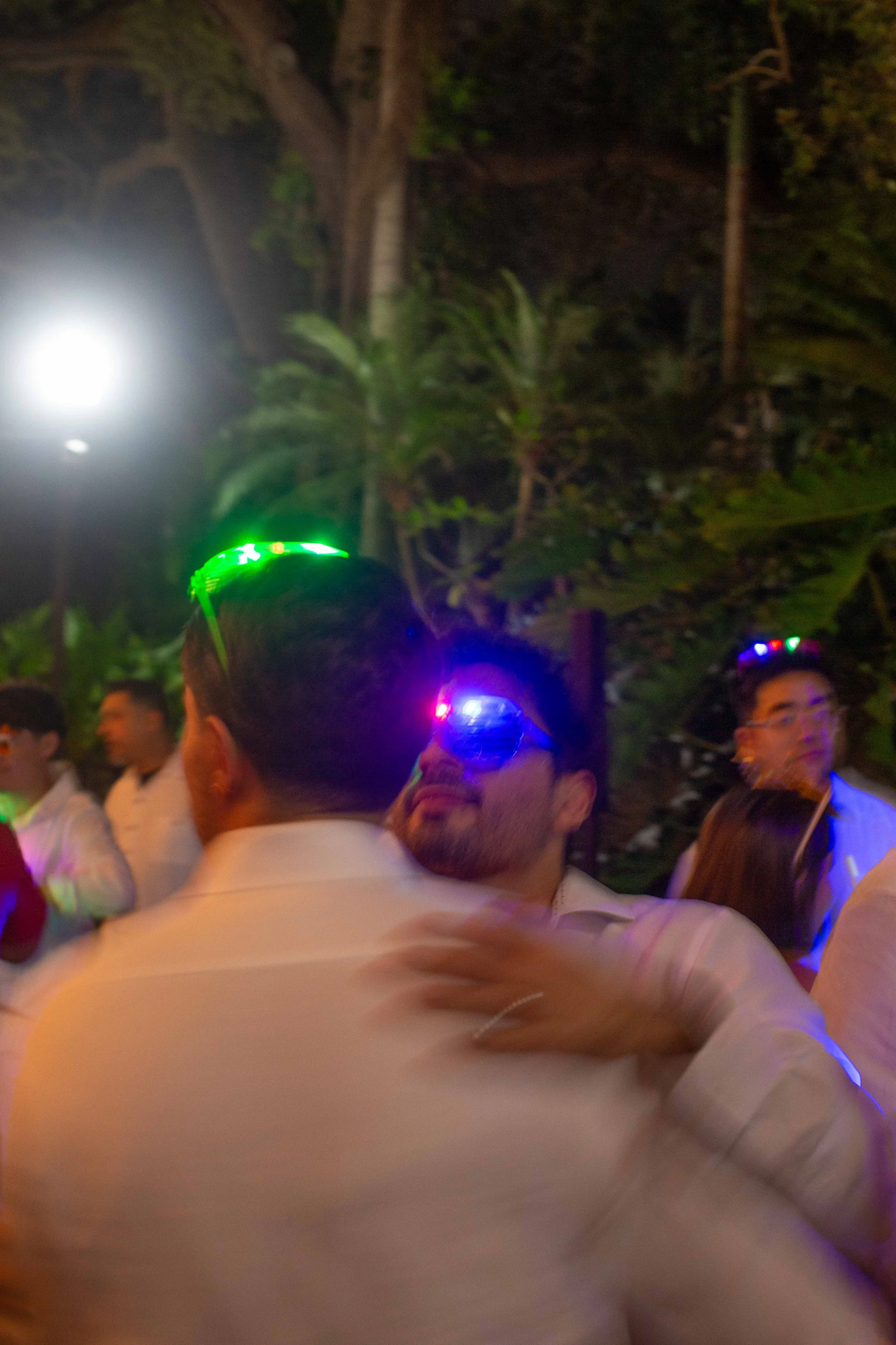 People wearing glow-in-the-dark glasses, dancing or socializing outdoors at night, with trees in the background.