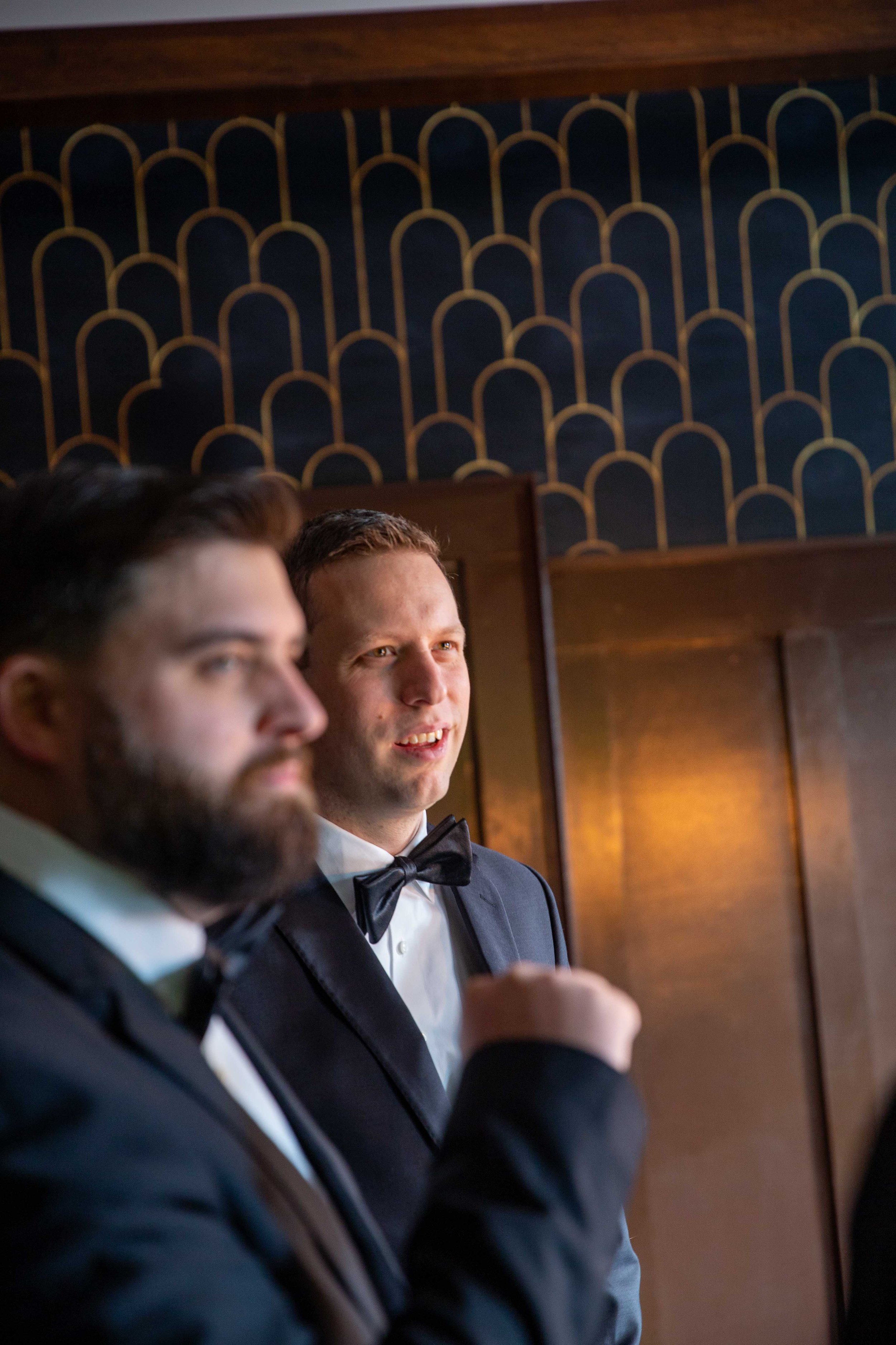 Two men in tuxedos, one with a beard and the other clean-shaven, standing indoors against a decorative wall with a geometric pattern, dressed for a formal event.