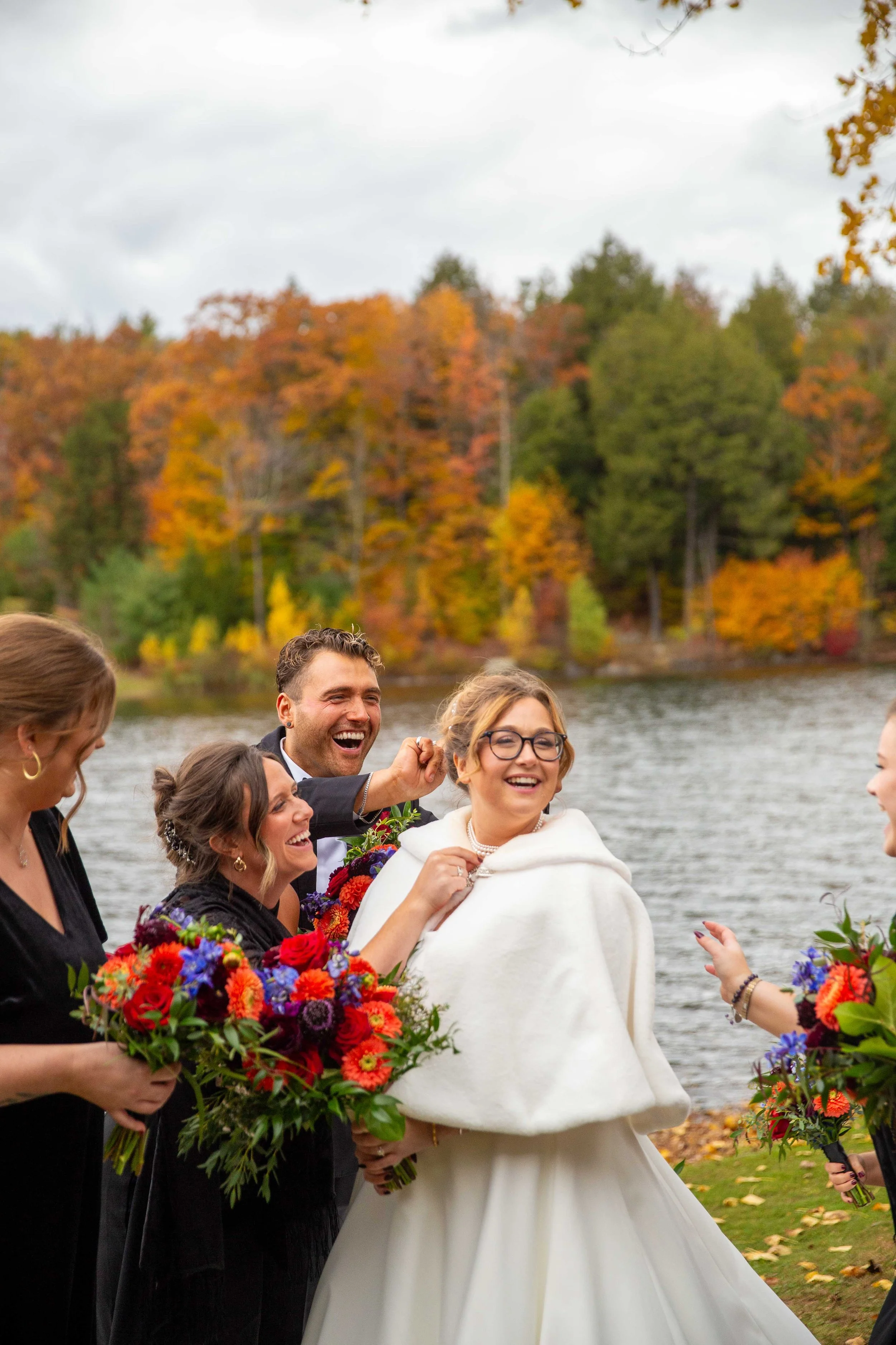 Happy group of people celebrating outdoors near a lake with fall trees in the background.