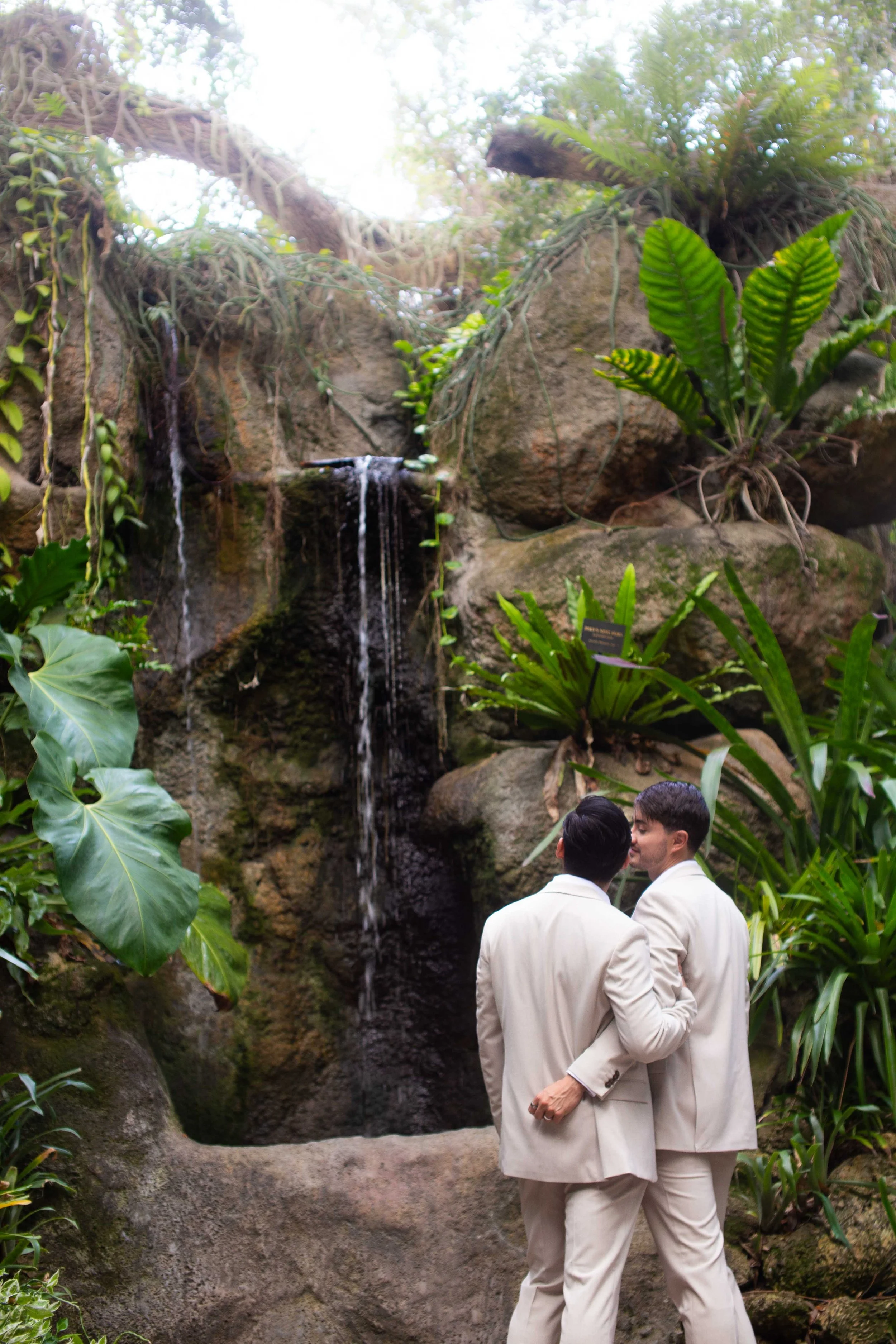 Two men dressed in white suits near a waterfall surrounded by lush green plants in a conservatory or botanical garden.