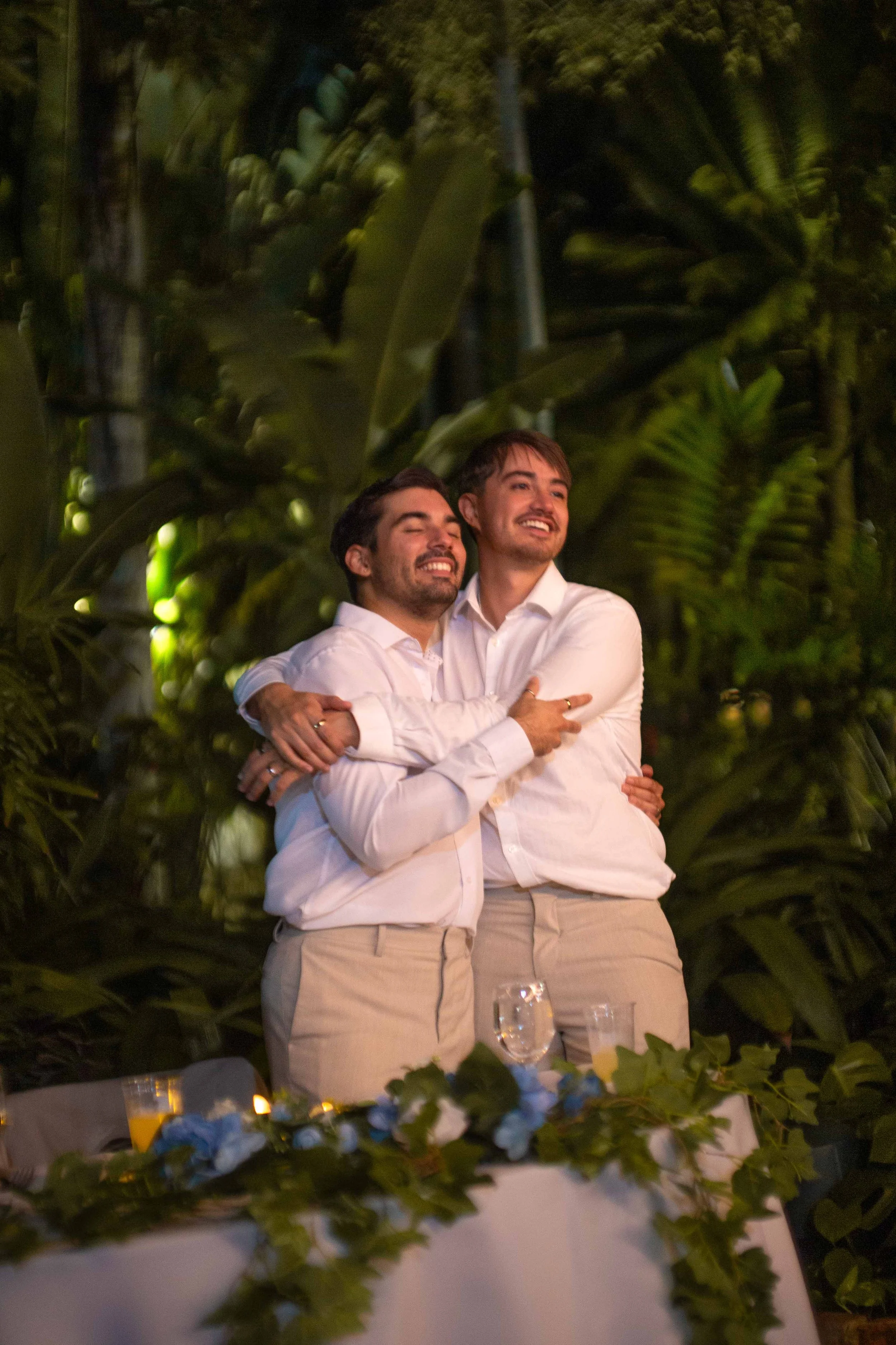 Two men embracing and smiling at an outdoor event at night, with dense green foliage in the background and a table with drinks and floral decorations in the foreground.