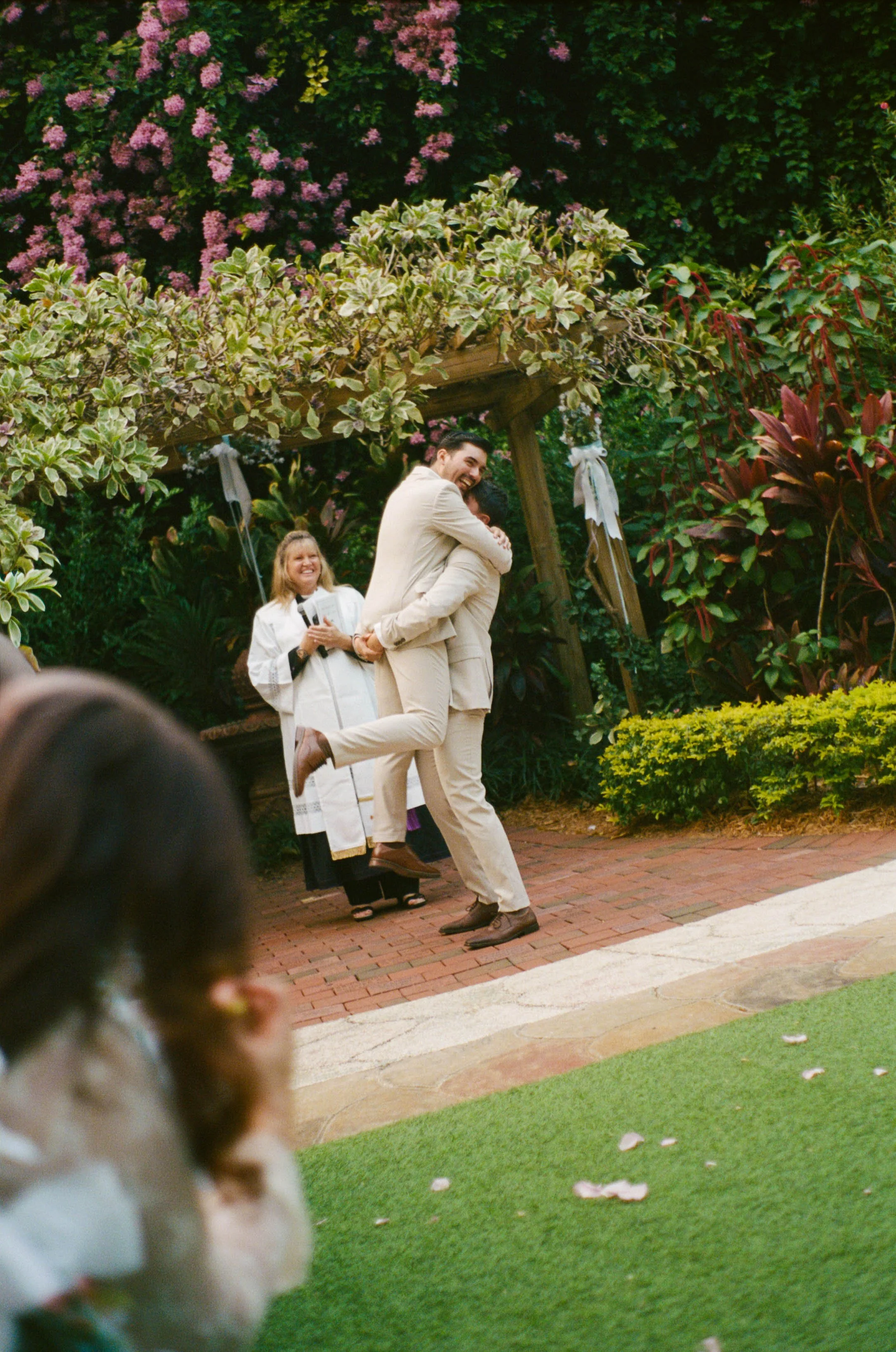 Two men in beige suits embracing, one lifting the other in celebration, with a woman dressed as a clergy member holding a microphone and smiling in the background, surrounded by greenery and pink flowers.