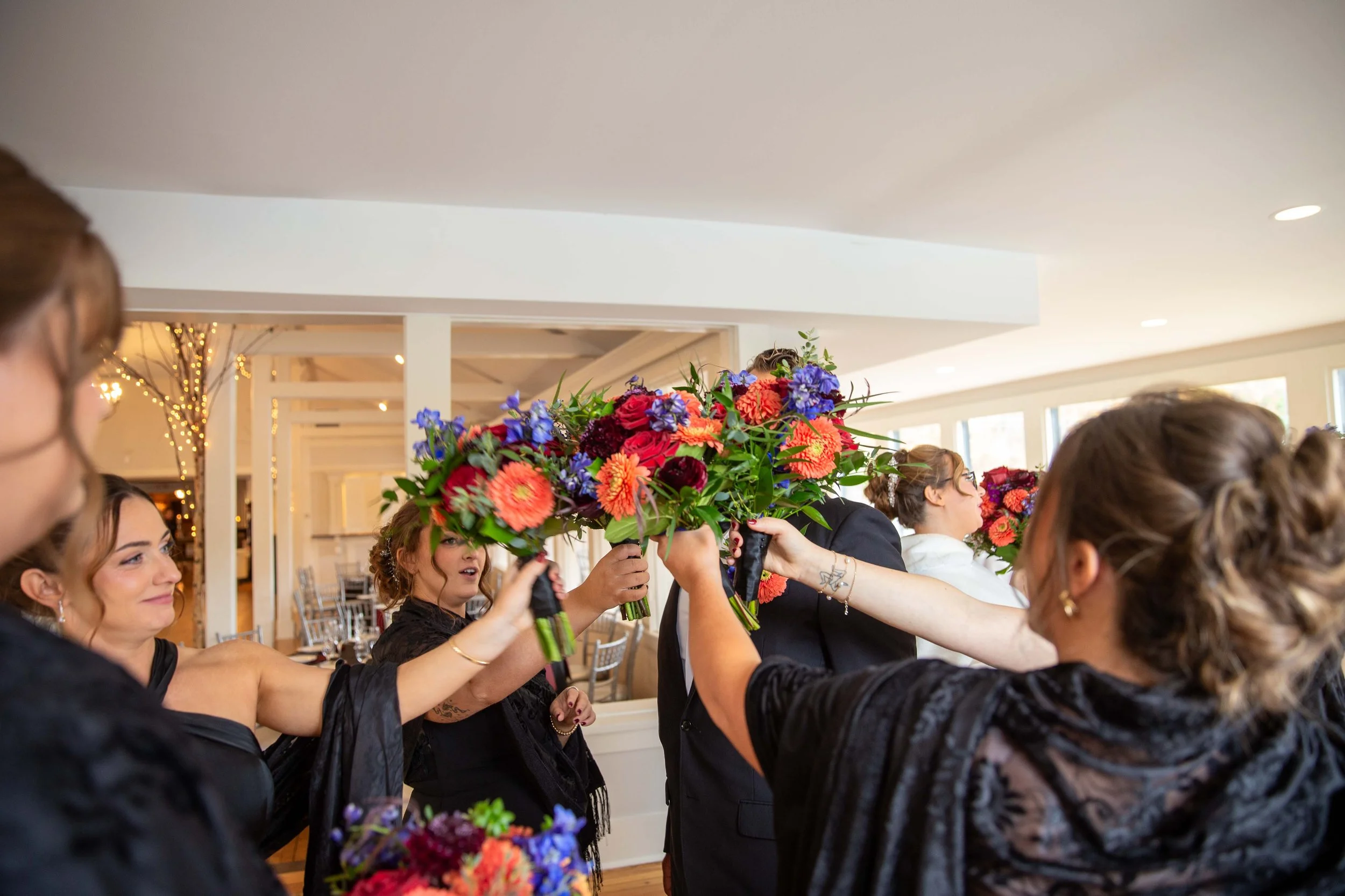 Group of women in black attire holding colorful flower bouquets in a celebration or wedding event inside a bright, decorated venue.