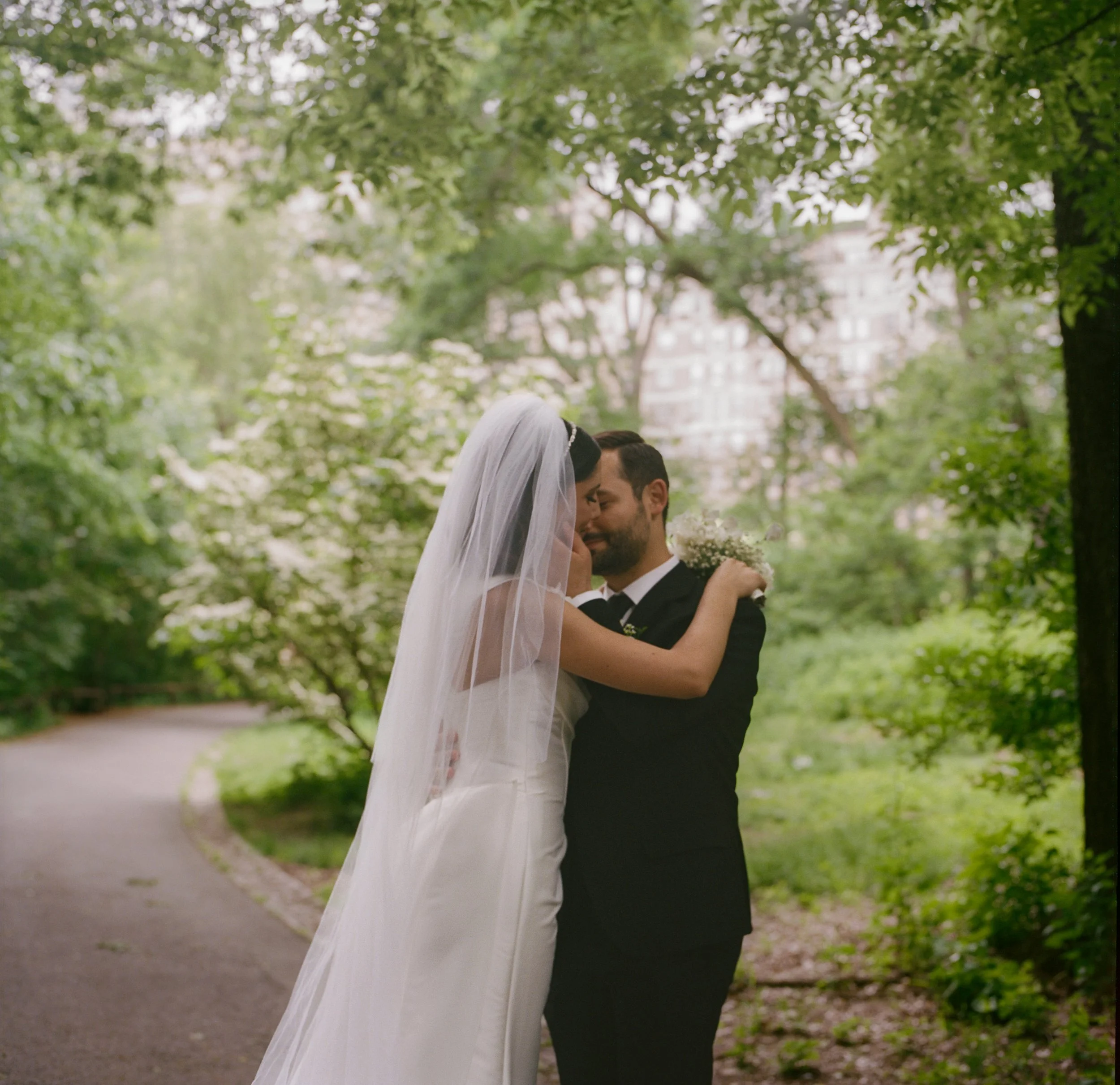 Bride and groom embracing in a lush green park, with trees and a winding path in the background.