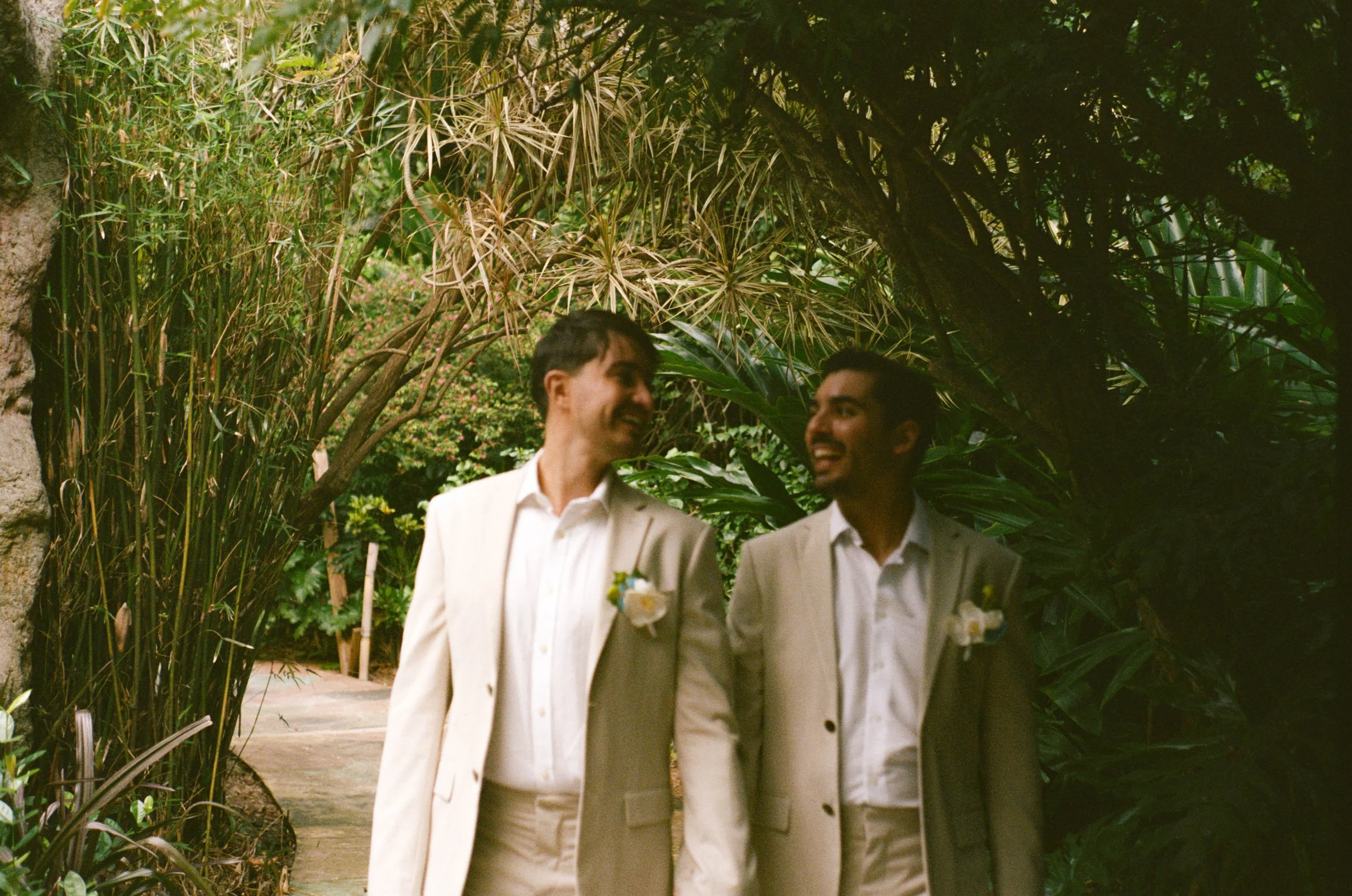 Two men in light-colored suits walking outdoors among lush green foliage, smiling and looking at each other.