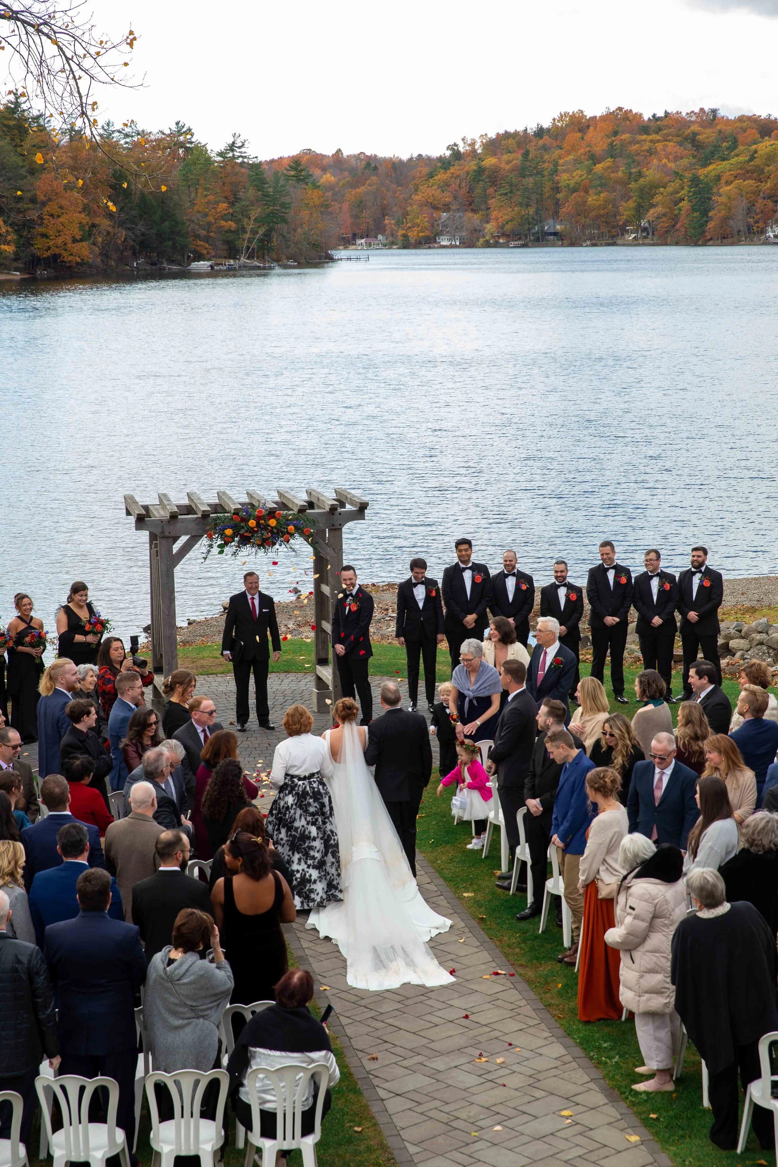 A wedding ceremony taking place outdoors by a lake with a backdrop of autumn trees. The bride and groom are walking down the aisle, surrounded by guests. The ceremony is officiated under a wooden arbor decorated with flowers.
