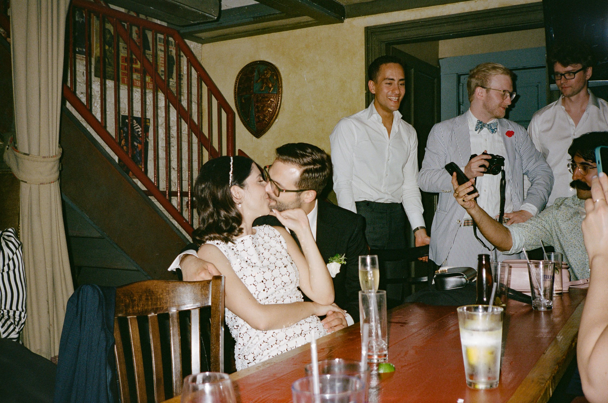 A group of people at a celebration, two are kissing at a table, while others stand around taking photos and smiling. The setting appears to be a cozy restaurant or tavern with a staircase and decorative shield on the wall in the background.