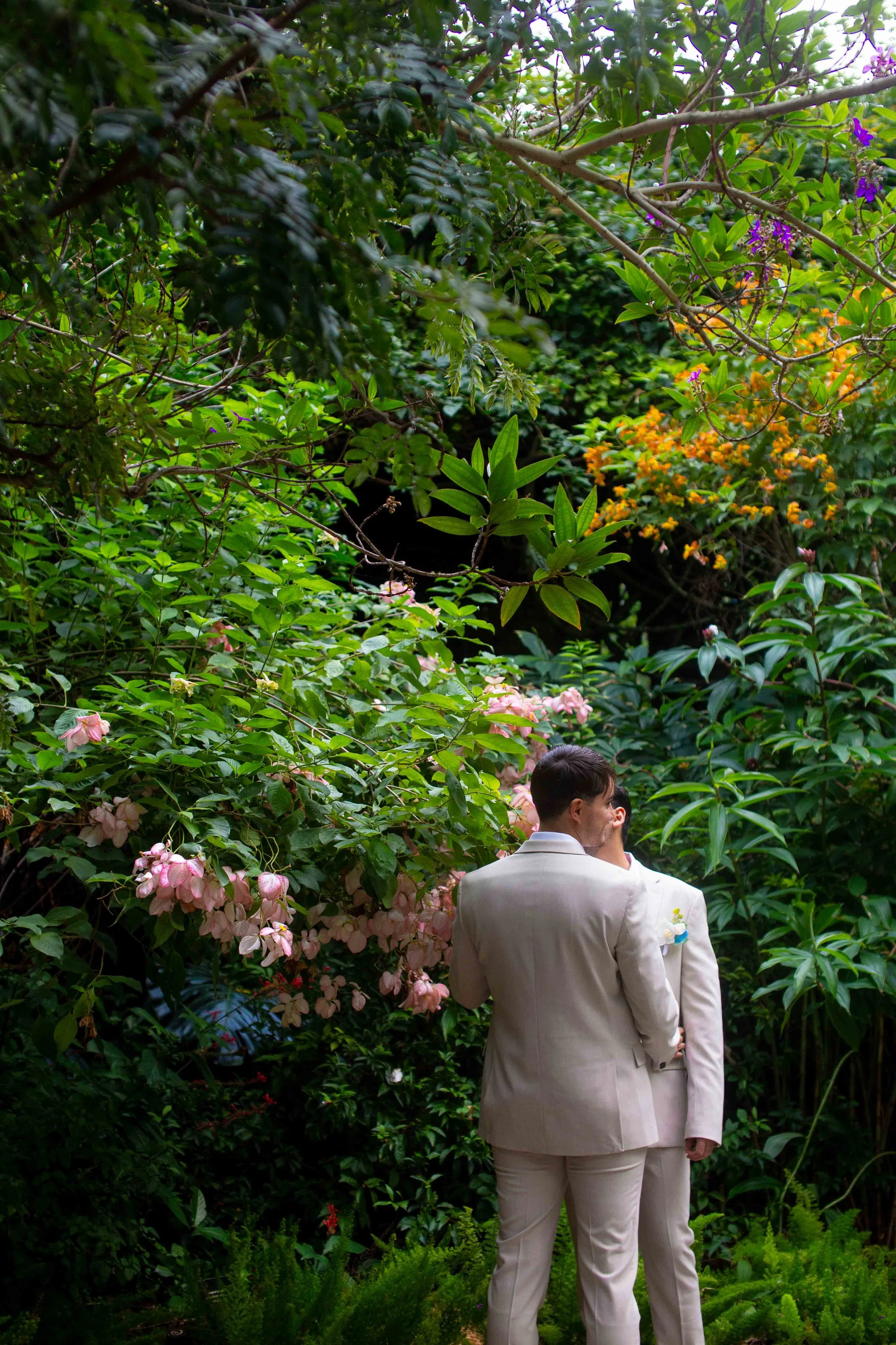 A couple in white suits embracing surrounded by lush green garden with pink and purple flowers.