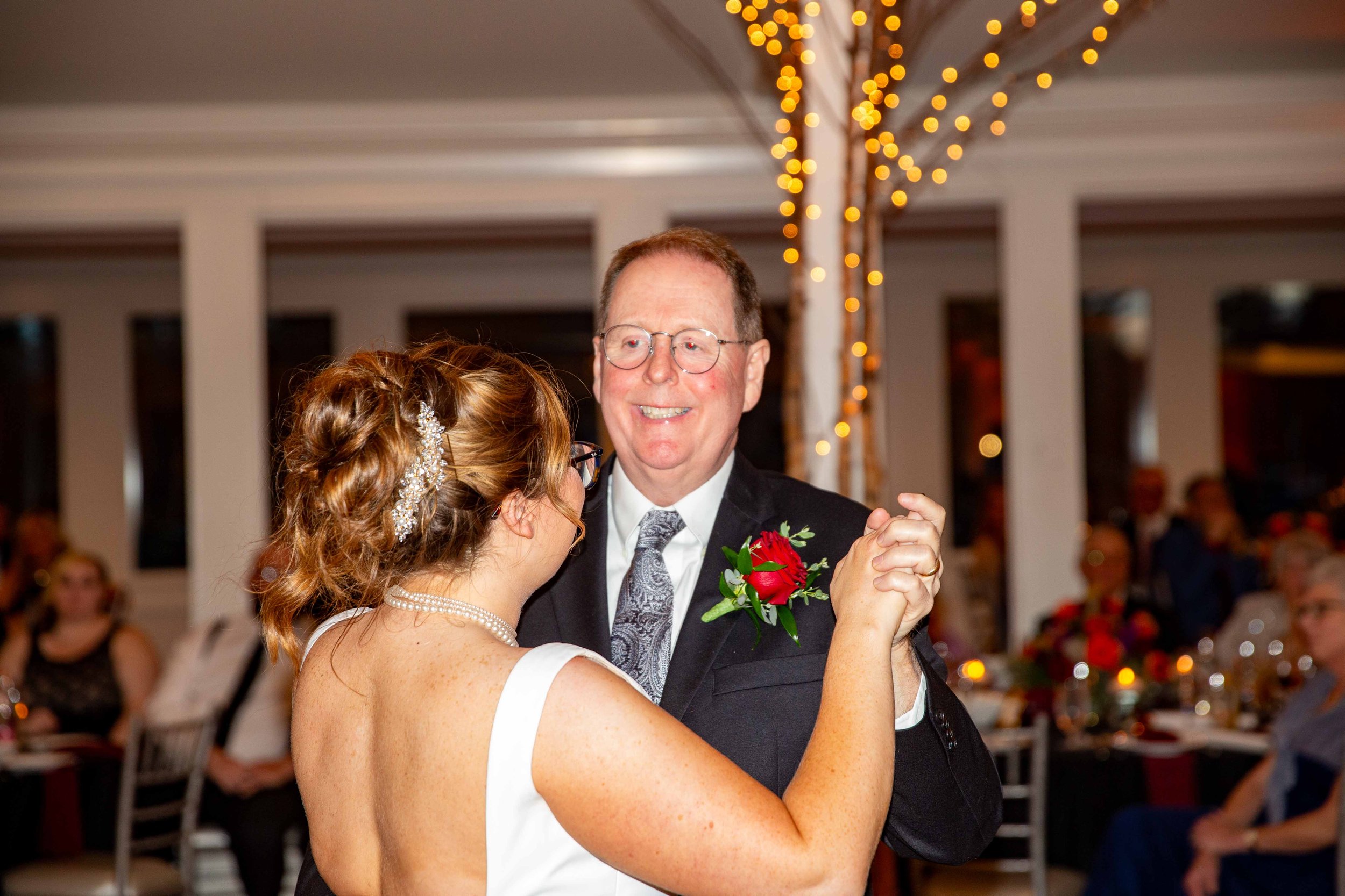 A bride and an older man dancing together at a wedding reception, with the bride wearing a white dress and pearl necklace, and the man in a suit with a red boutonniere, in a decorated reception hall with string lights and guests in the background.