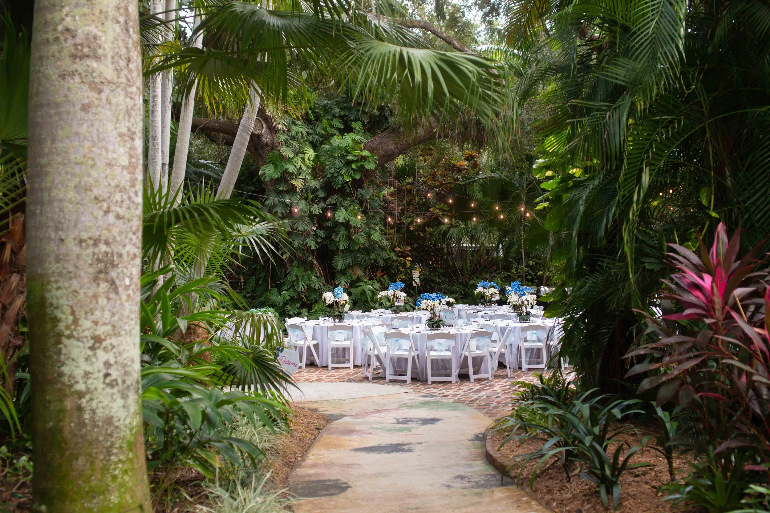 Outdoor dining area decorated with white tablecloths and floral centerpieces, surrounded by lush green tropical plants and trees, with string lights overhead.