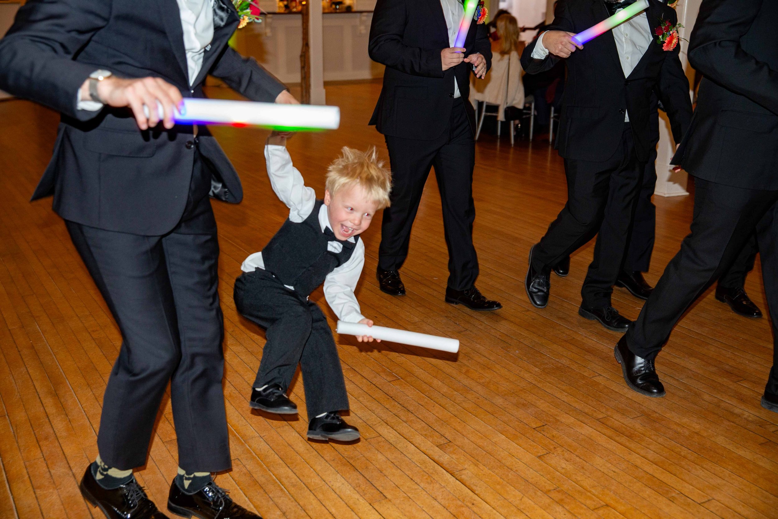 Young boy in formal attire enjoying a lively dance with adults, all holding glow sticks, on a polished wooden floor at a celebration or party.