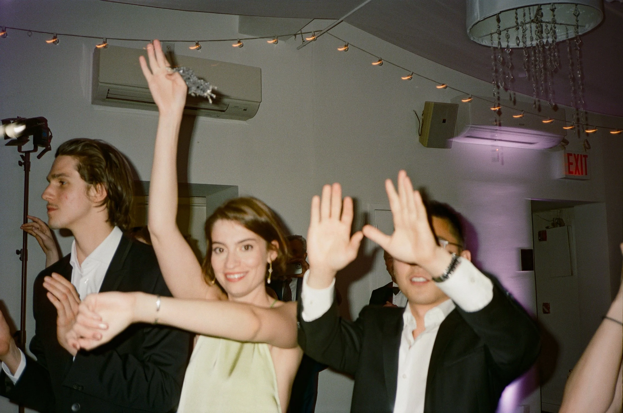People dancing at a party, woman smiling and waving, man in suit with his hand raised, women and men in formal attire, festive lighting, and decorations in the background.