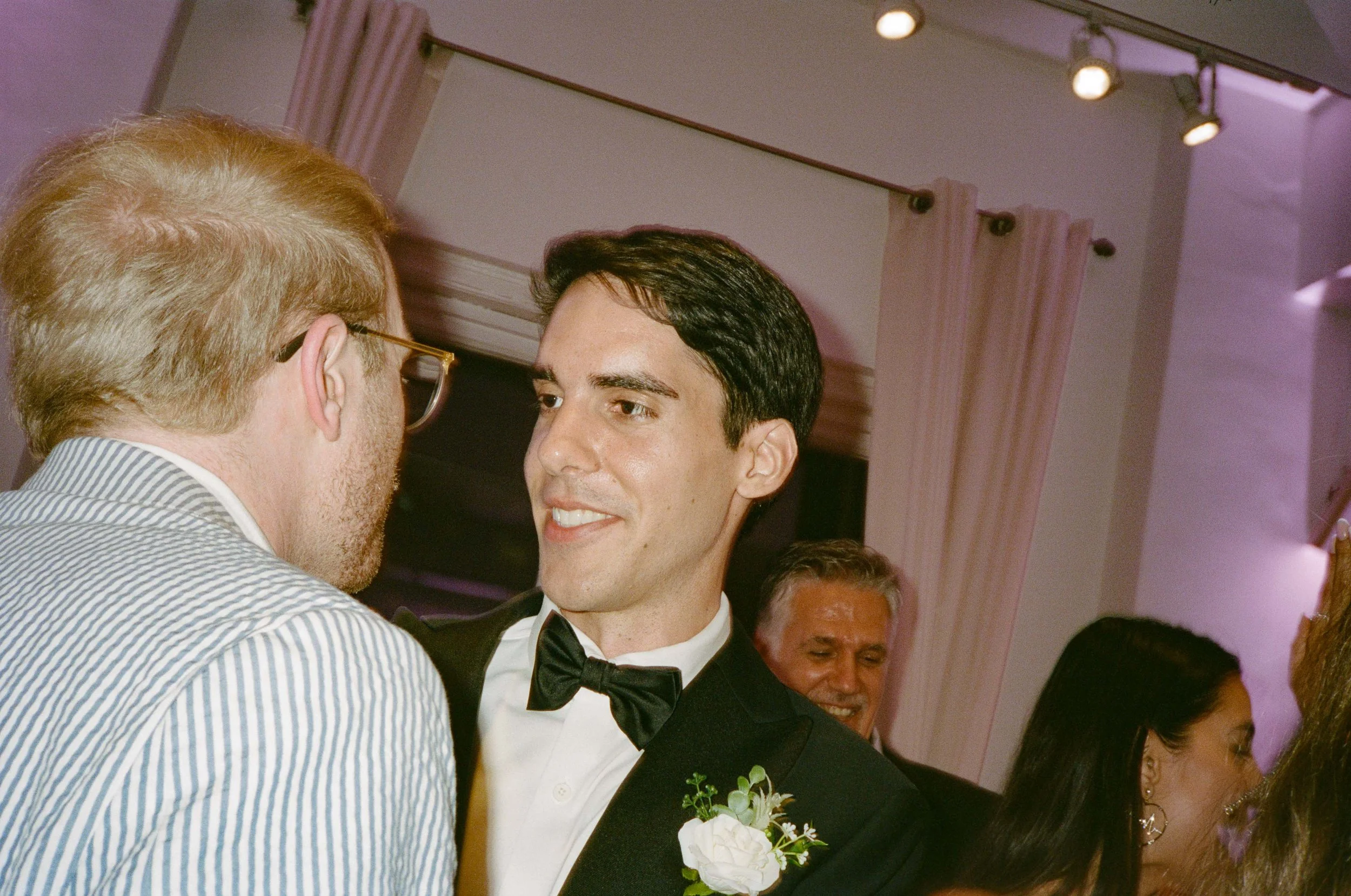 Two men engaged in conversation at a formal event, one wearing a tuxedo with a boutonniere, and the other in a striped shirt with glasses, with several other people in the background.