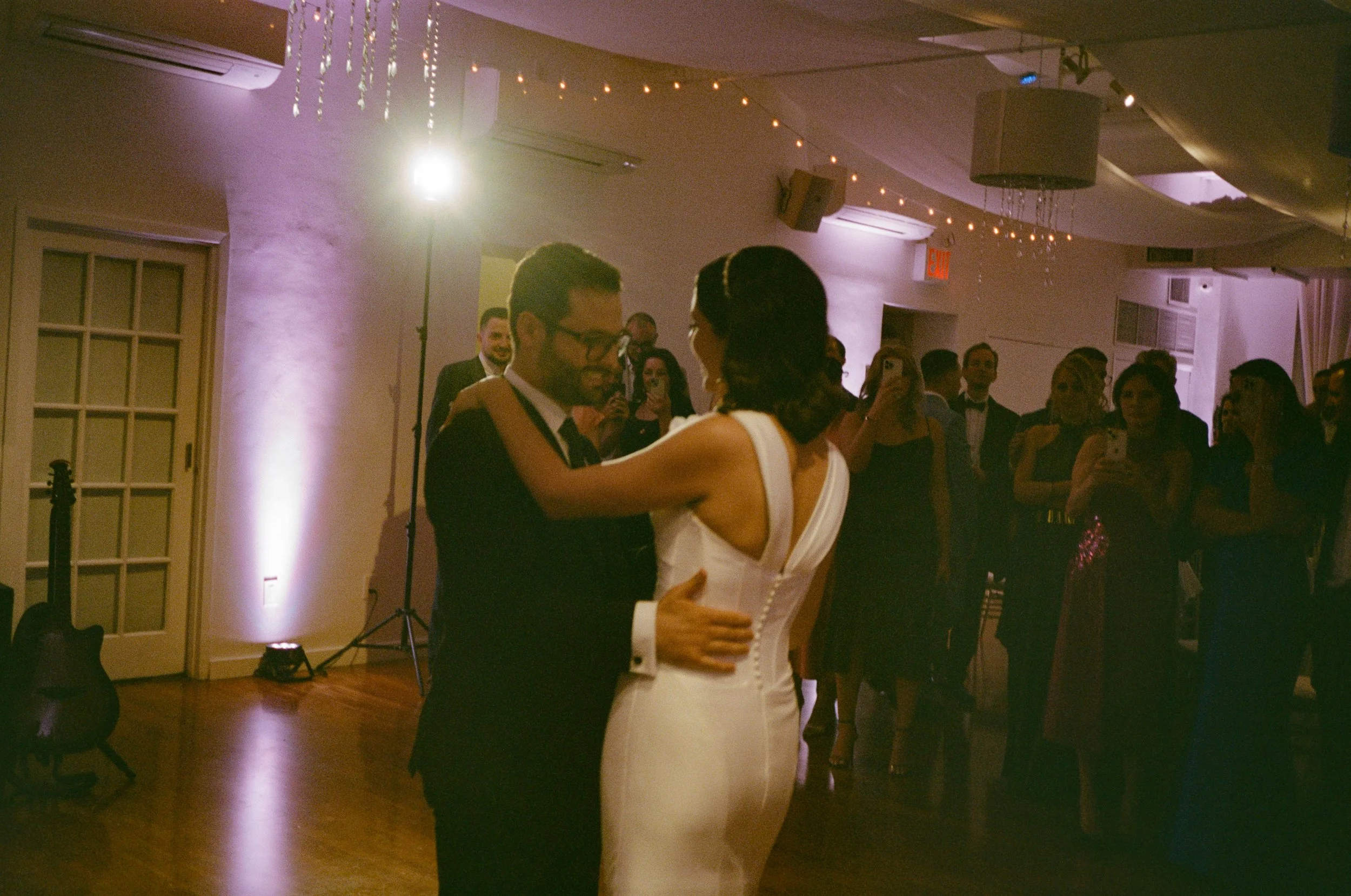 A bride and groom dance together at their wedding reception with guests watching and taking photos in the background.