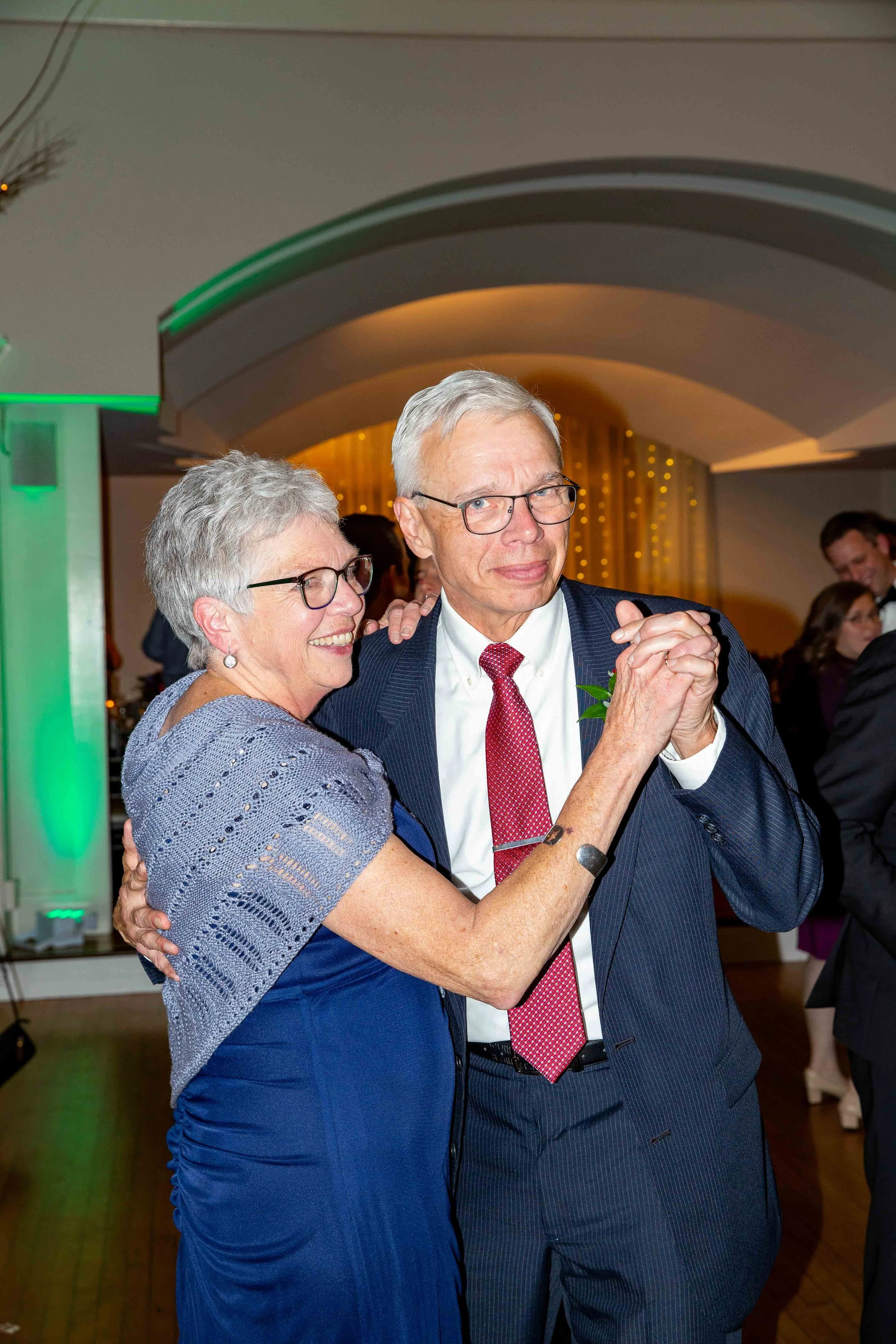 An elderly couple dancing together at a formal event, with the woman smiling and wearing a blue dress with an openwork shawl, and the man in a dark suit with a red tie, smiling slightly, in a decorated venue with warm lighting.