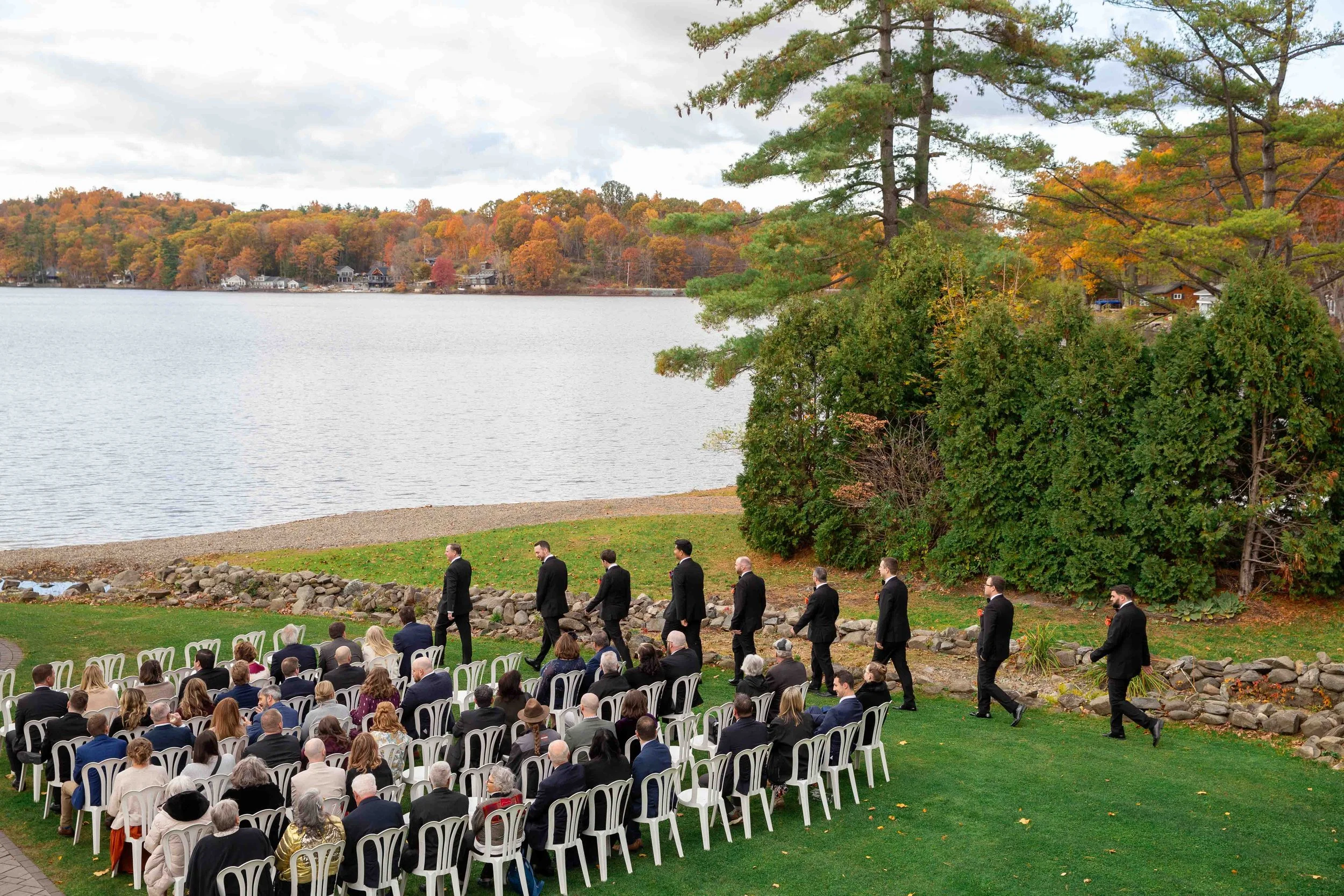 Outdoor wedding ceremony by a lake, with guests seated in white chairs and groomsmen in black suits walking past.