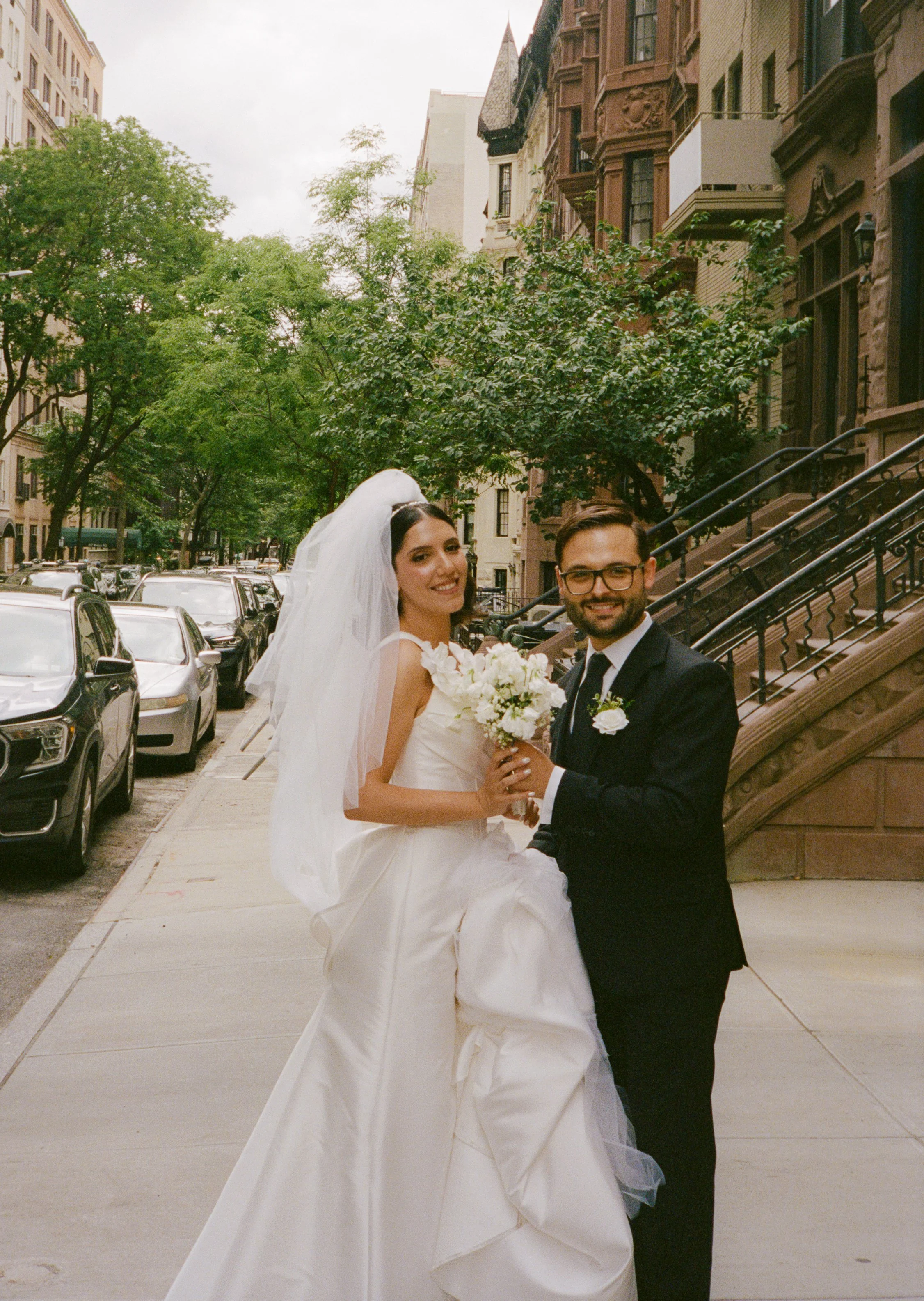 A bride and groom pose on a city sidewalk after exchanging vows, with trees and brownstone buildings in the background.