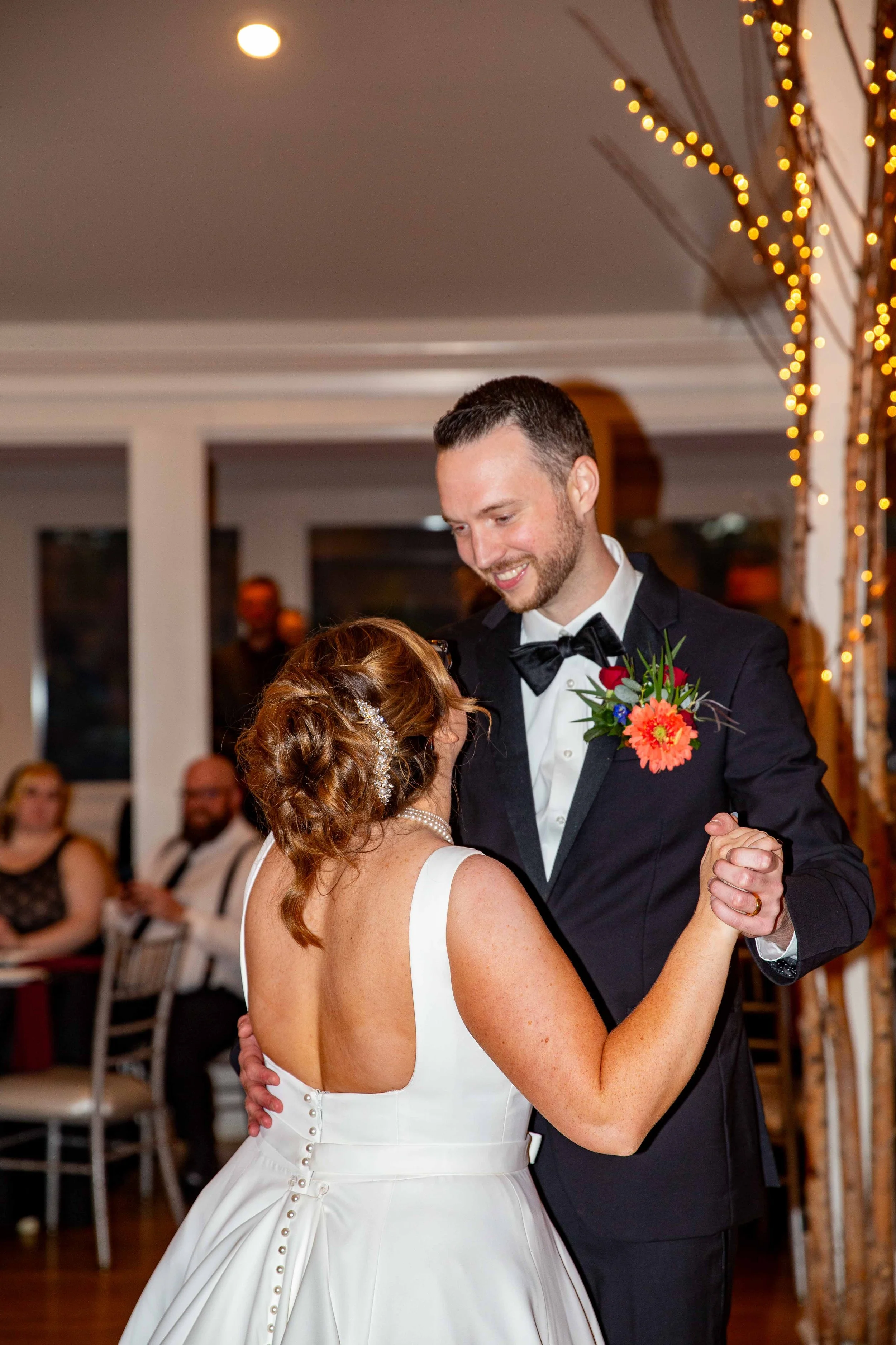 A bride and groom dancing at their wedding reception, with the groom smiling and wearing a tuxedo with a flower boutonniere, and the bride in a white wedding dress with pearl accents, in a decorated venue with string lights.
