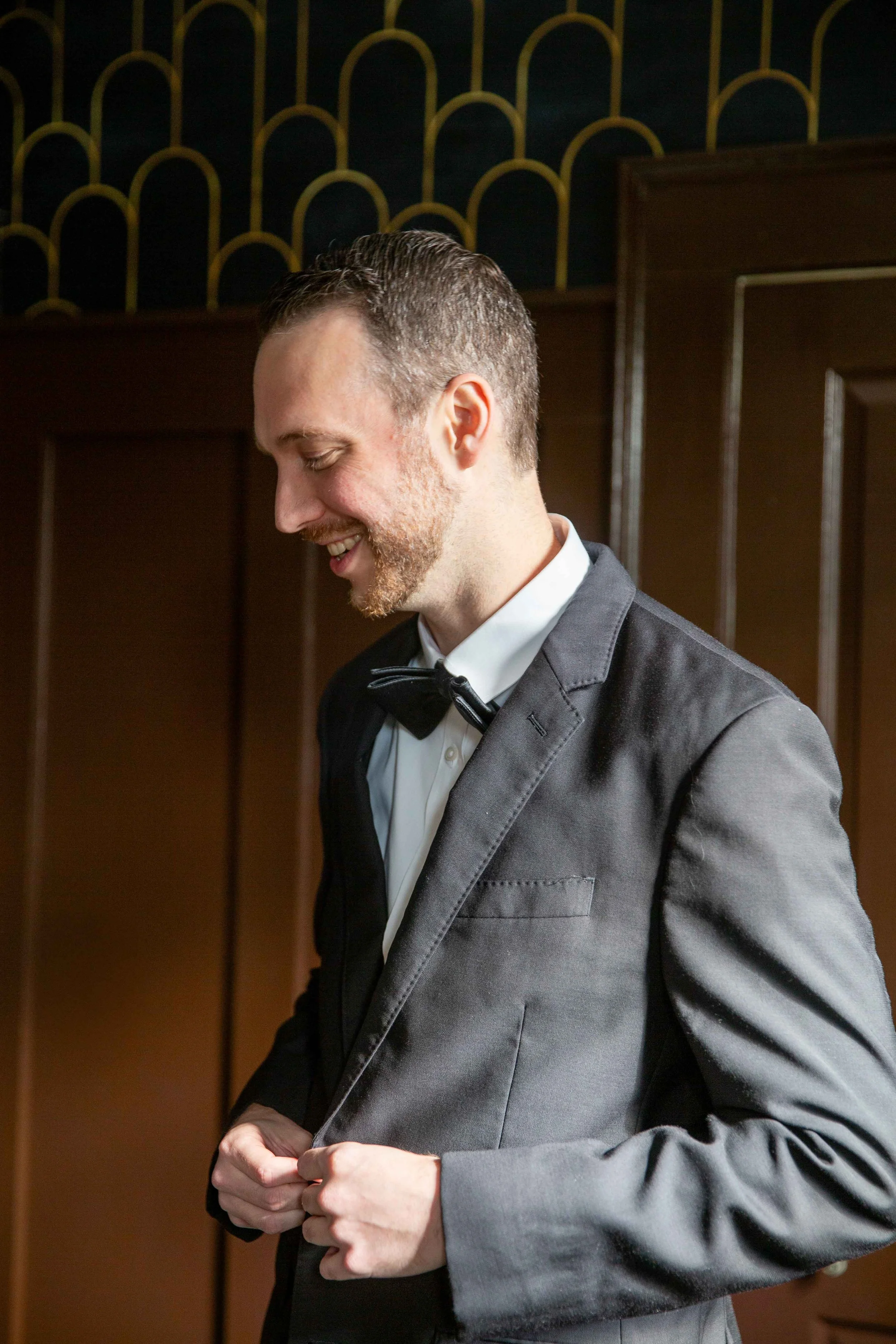 A man in a tuxedo adjusting his jacket, smiling, with a patterned wall and wooden paneling in the background.